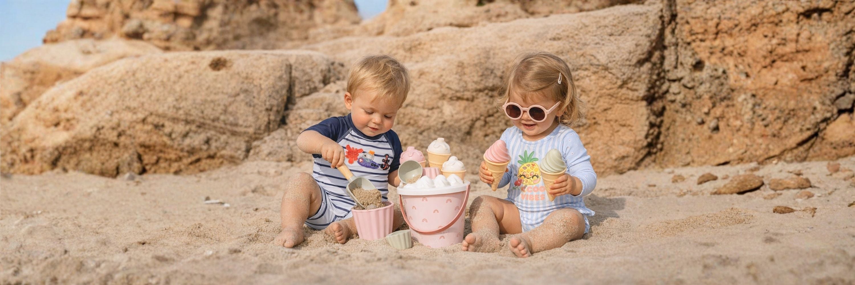 Two toddlers sit on the beach playing with sand toys, smiling and enjoying a sunny day by the rocks.