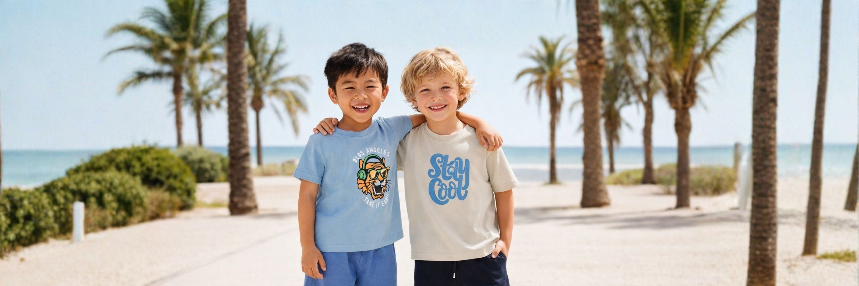 Two boys smiling on a beach path with arms around each other, wearing casual T-shirts—one with a graphic print and the other with “Stay Cool,” with palm trees and sea in the background.