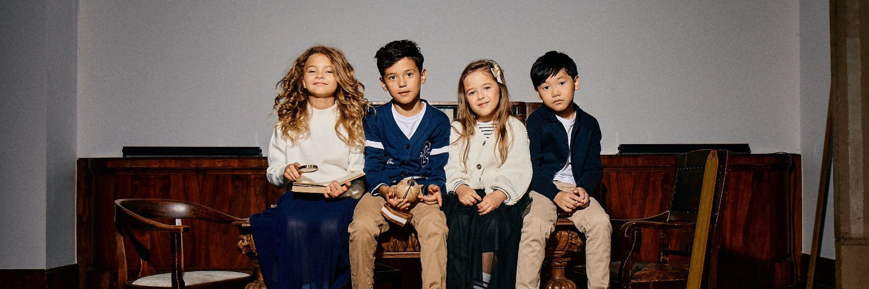 Four children in elegant clothes are sitting side by side in a stylized interior, holding a small globe, a book, and a magnifying glass.