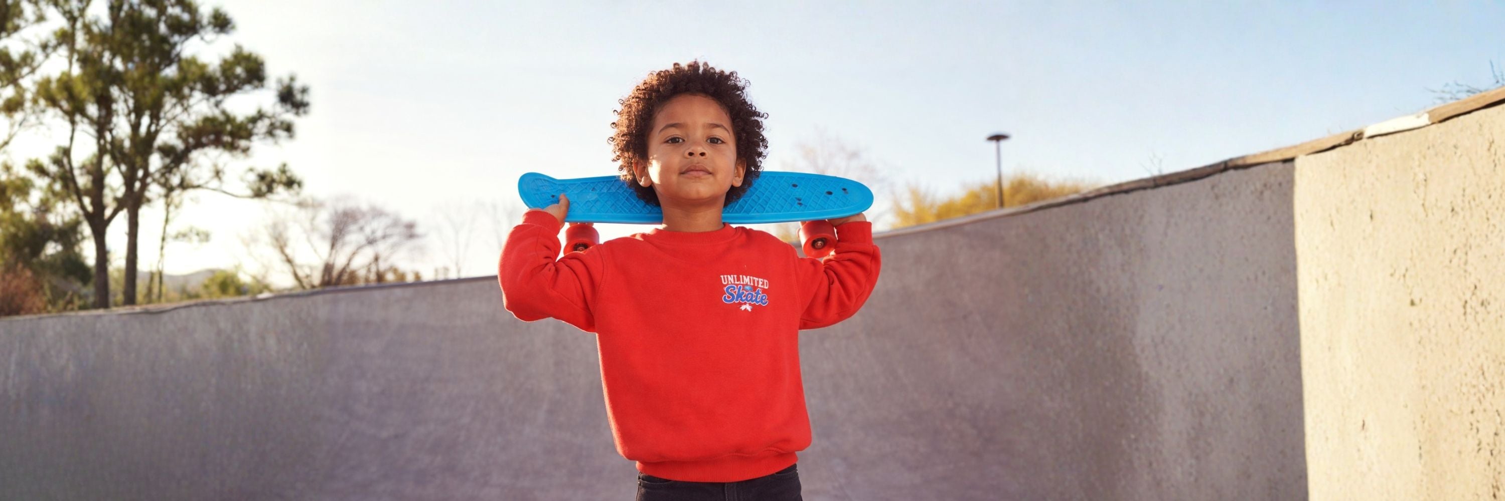 A boy holding a skateboard over his shoulders, wearing a red sweatshirt, standing outdoors in a skate park with trees and a clear sky in the background.