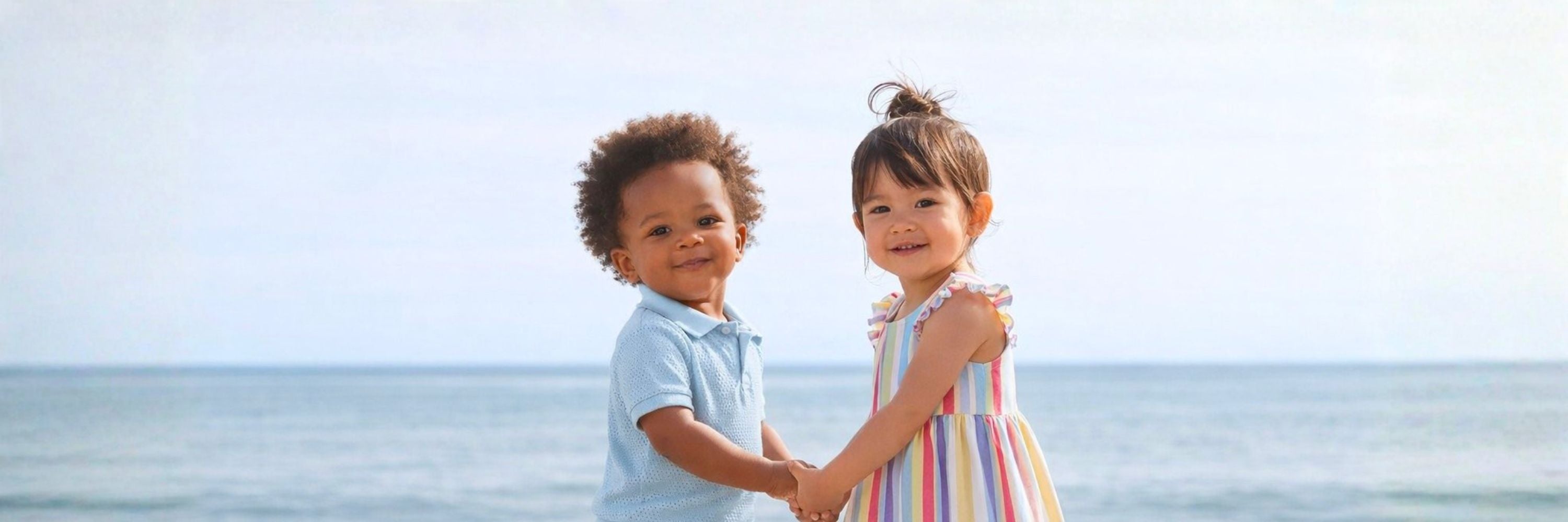 Two toddlers holding hands at the beach, smiling at the camera, with the ocean in the background.