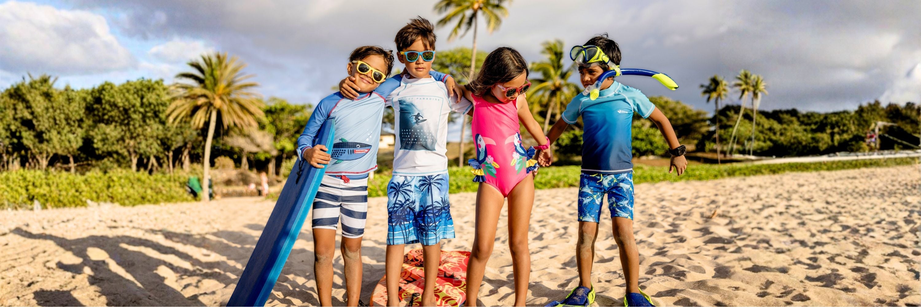 Four kids posing together on a sandy beach. They are wearing UV rash guards, swim shorts, and a pink swimsuit with a toucan print. Some children have sunglasses, one holds a bodyboard, and another wears a snorkeling mask and fins.