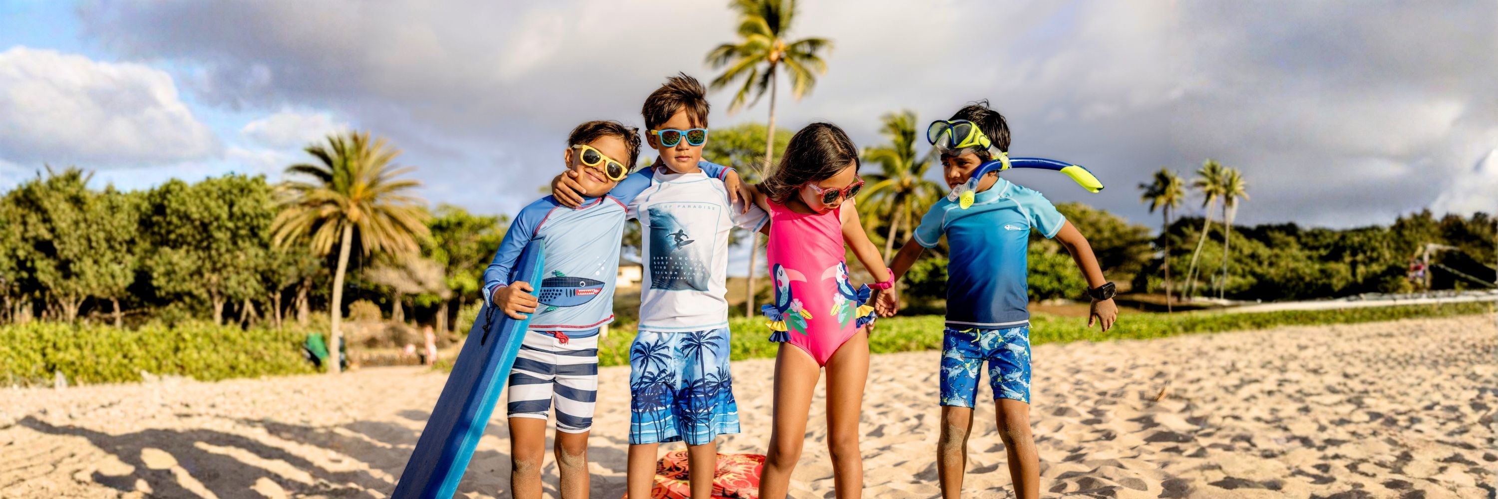 Four children standing on a sandy beach wearing colorful swimwear and sunglasses, with one child wearing a snorkel mask.