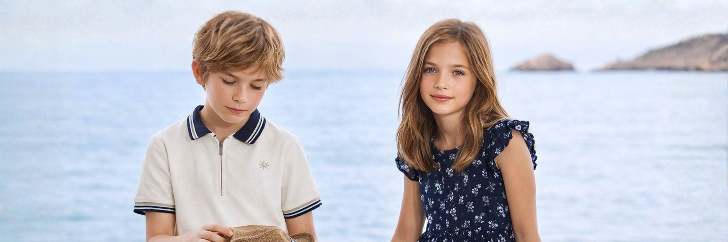 Two children standing by the seaside, a boy in a light polo shirt looking down and a girl in a dark floral dress facing the camera, with the ocean and distant coastline in the background.