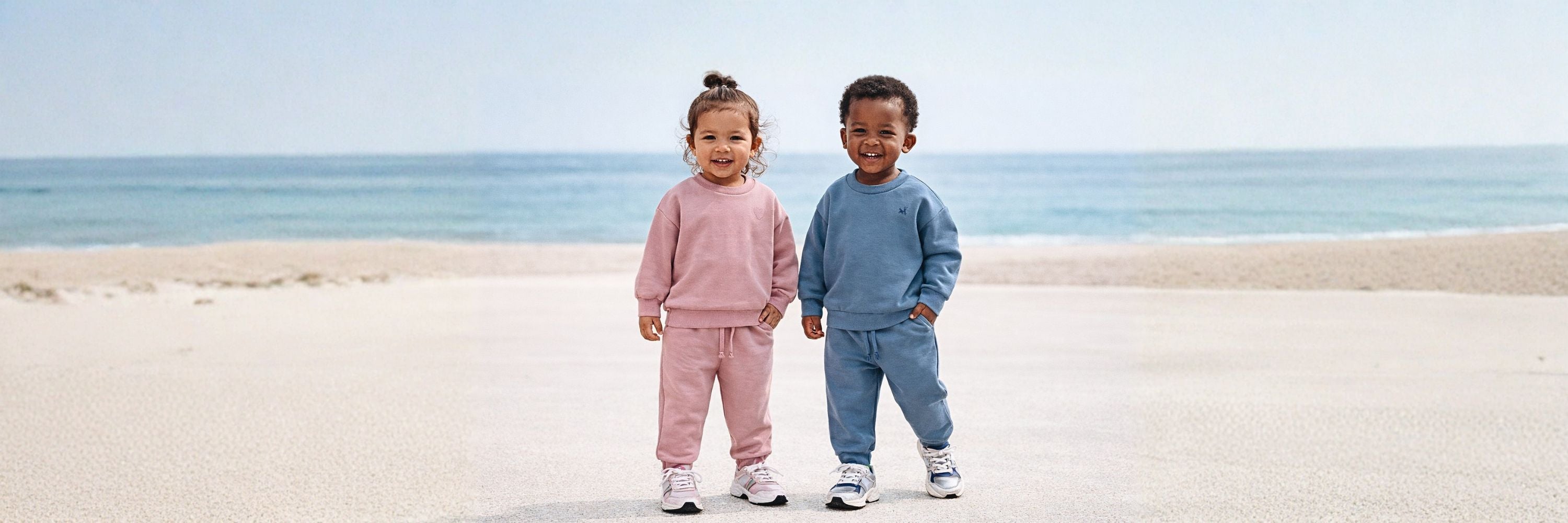 Two toddlers standing on the beach, smiling at the camera, wearing matching tracksuits in pink and blue, with the ocean in the background.