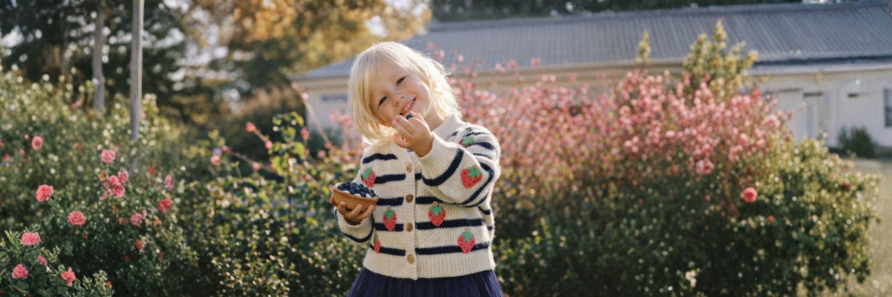 A smiling girl standing in a garden, wearing a striped cardigan with strawberry details, holding and eating berries with flowers and greenery in the background.
