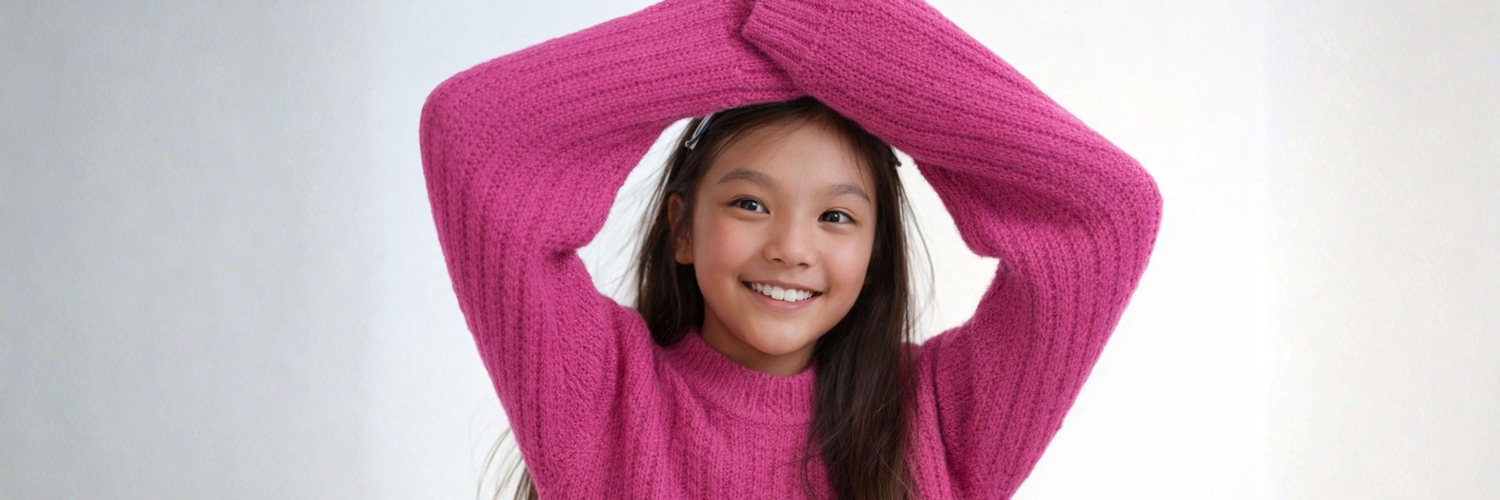 Smiling girl in a bright pink sweater raises her arms playfully, creating a cheerful and cozy look against a clean, minimal background.