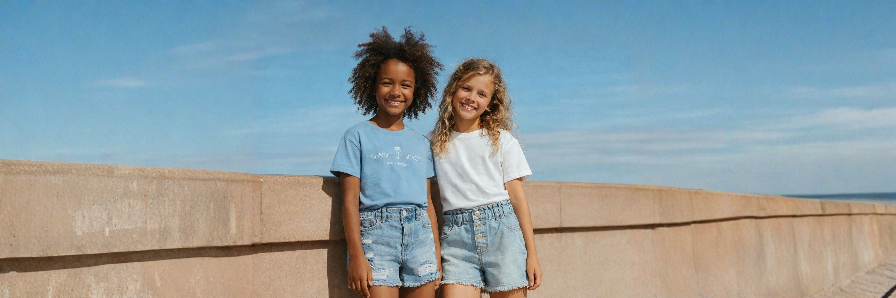 Two girls stand by a seaside wall in casual denim shorts and T-shirts, smiling in the warm sunlight with a relaxed, summery atmosphere.
