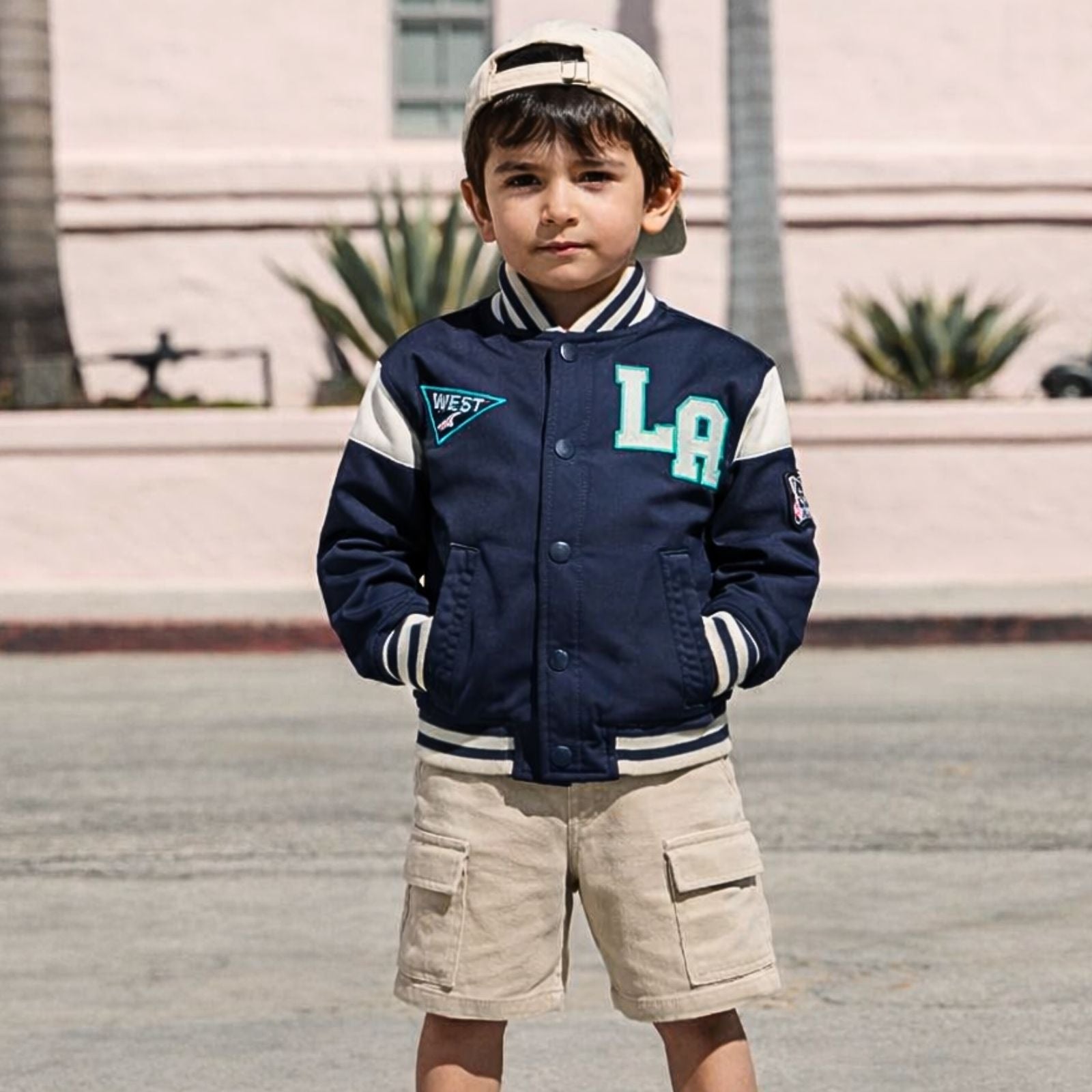 Child wearing a navy blue varsity jacket with 'LA' and 'West' lettering, standing outdoors.
