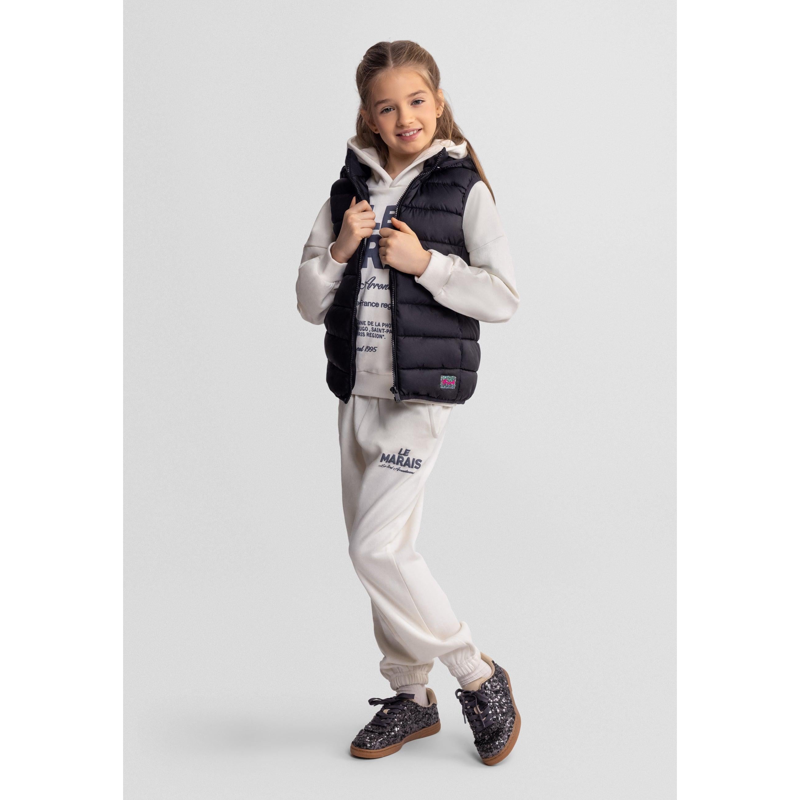 Girl in black quilted gilet zipped up over white hoodie and cream tracksuit, hands positioned on zip, against white studio background