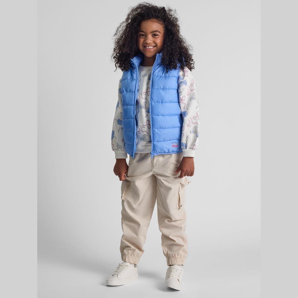 Young girl wearing light blue quilted gilet with detachable hood over patterned sweatshirt and beige cargo trousers, full body front view on white background