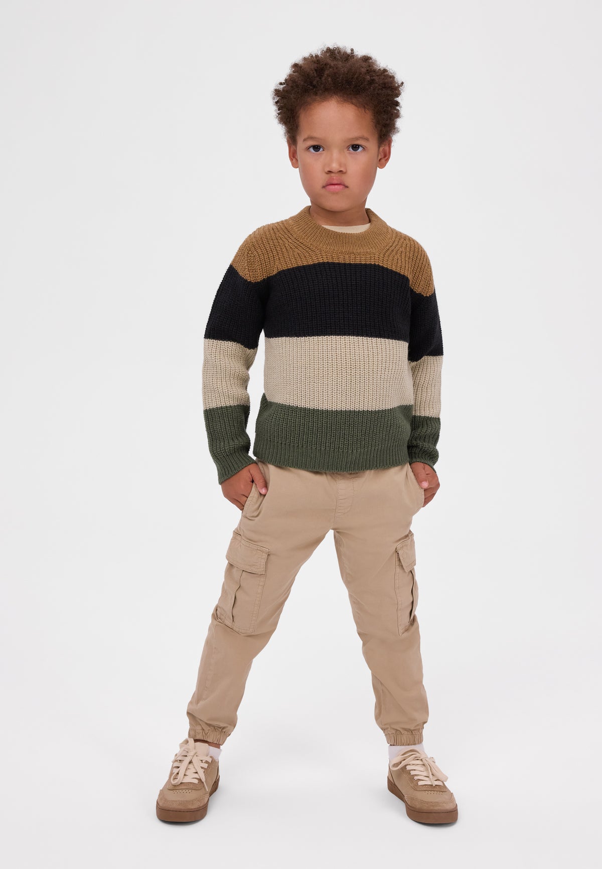A young child modeling a color-blocked knit sweater with horizontal stripes in brown, black, cream, and olive green, paired with beige cargo pants and tan sneakers, against a plain light gray studio background.