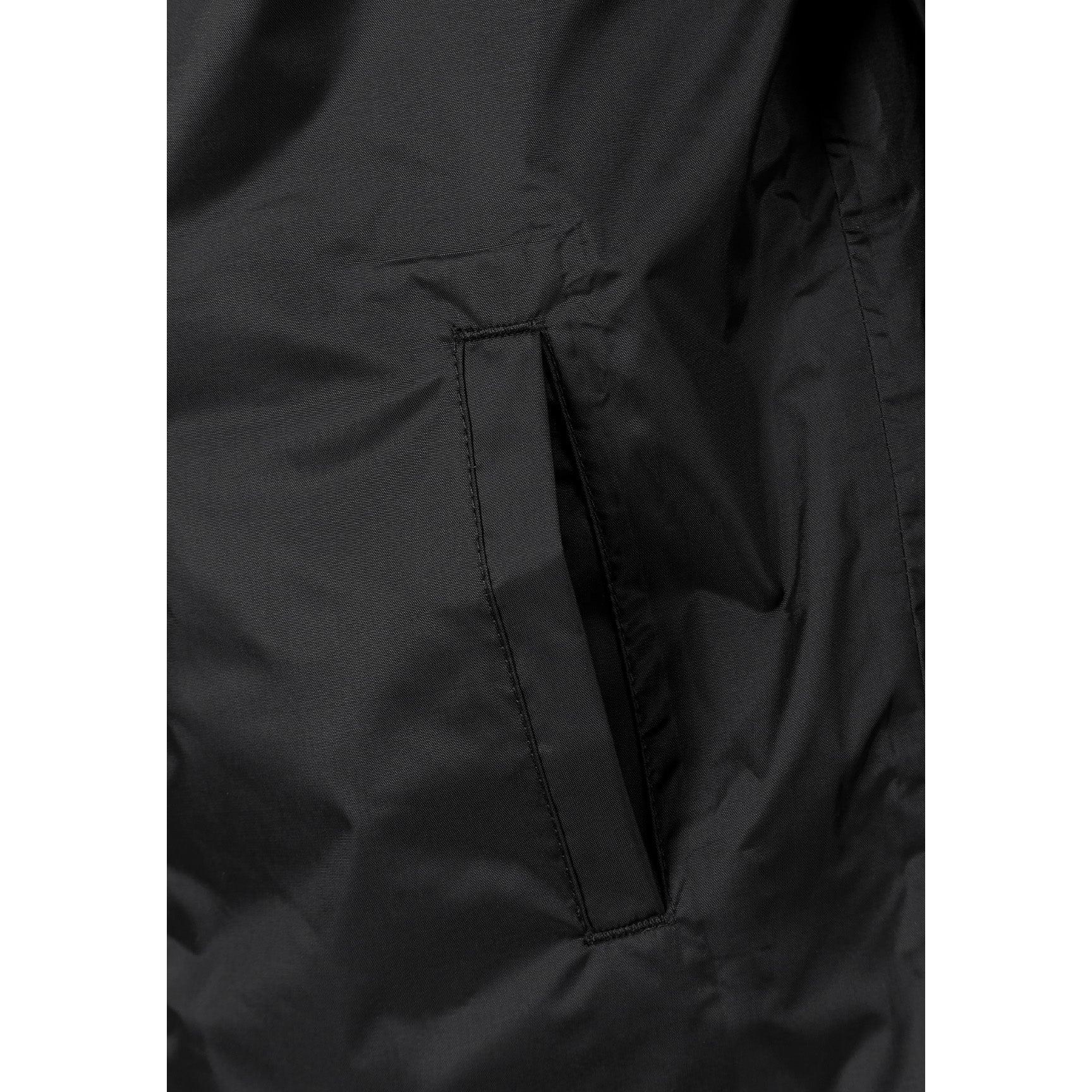 Boy in black hooded waterproof jacket pointing forward, wearing light blue cargo jeans and black sneakers on white studio background