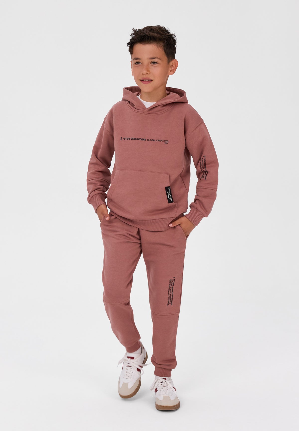 A young boy modeling a dusty rose pink two-piece tracksuit, featuring a pullover hoodie with black graphic text on the chest and sleeve, matching jogger pants with vertical text graphics, and white sneakers, set against a plain light gray studio background.