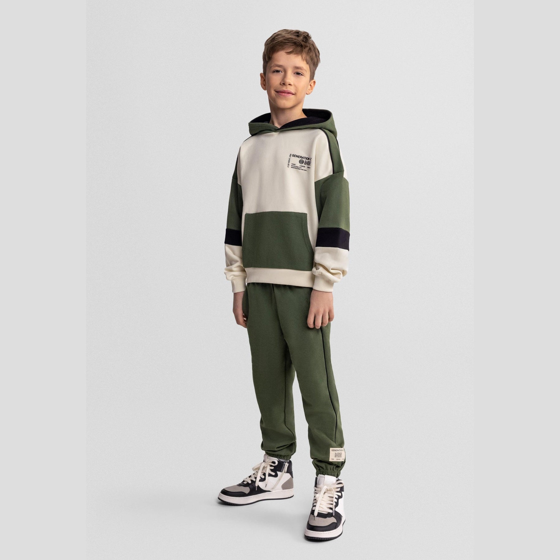 Boy wearing khaki tracksuit set with hooded sweatshirt and joggers against light background, full body front view