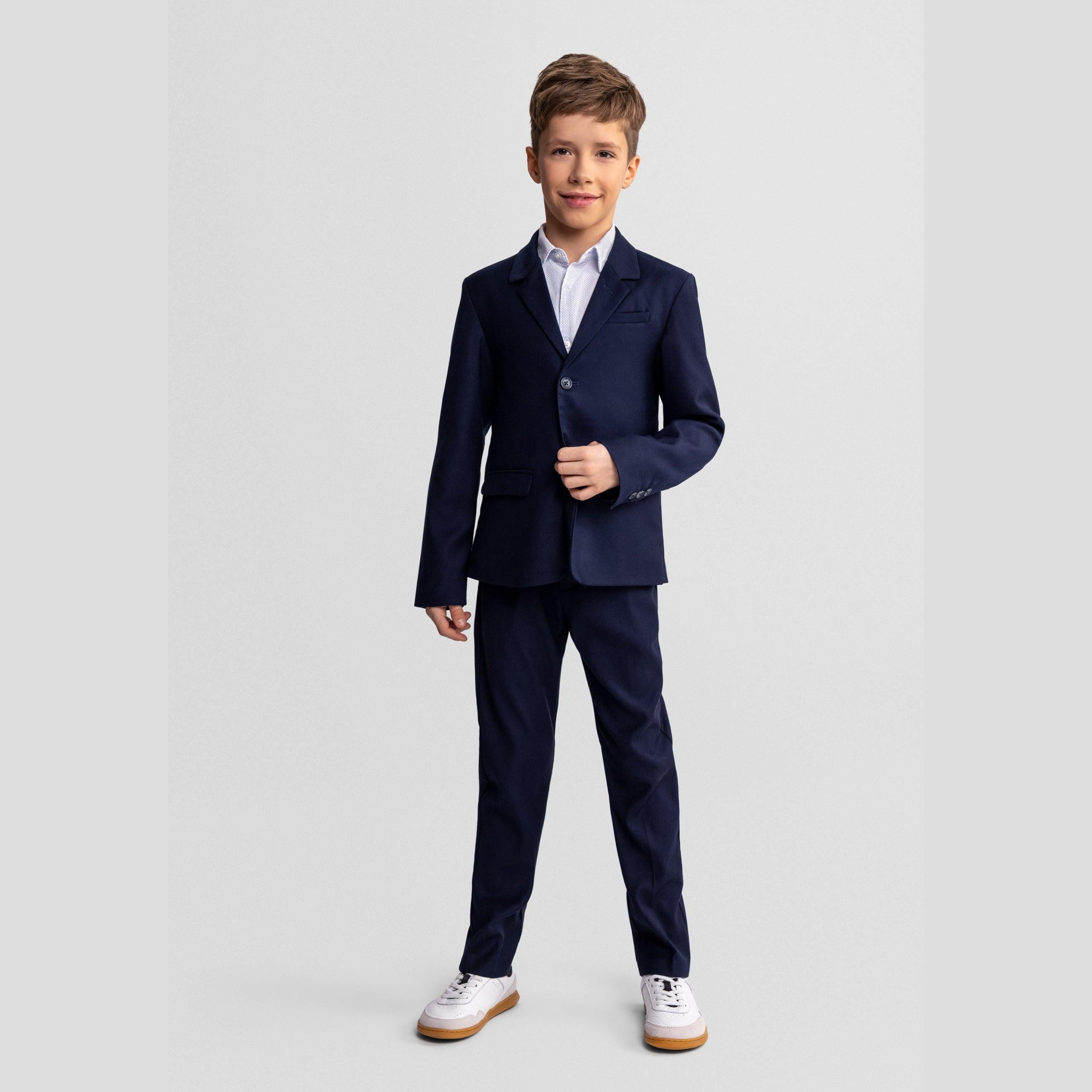 Young boy with curly hair wearing complete navy formal suit with waistcoat and tie, standing against light grey background