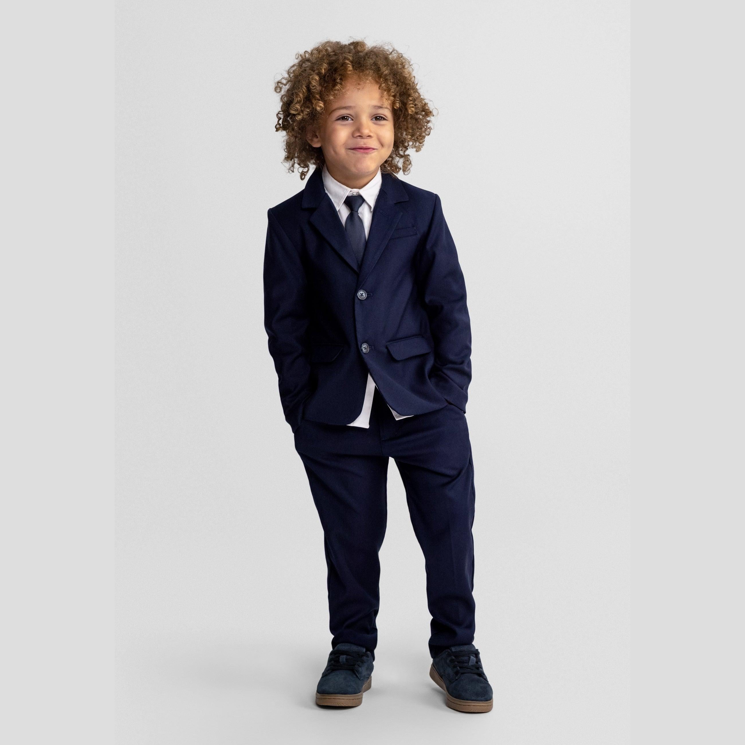 Young boy with curly hair wearing complete navy formal suit with waistcoat and tie, standing against light grey background
