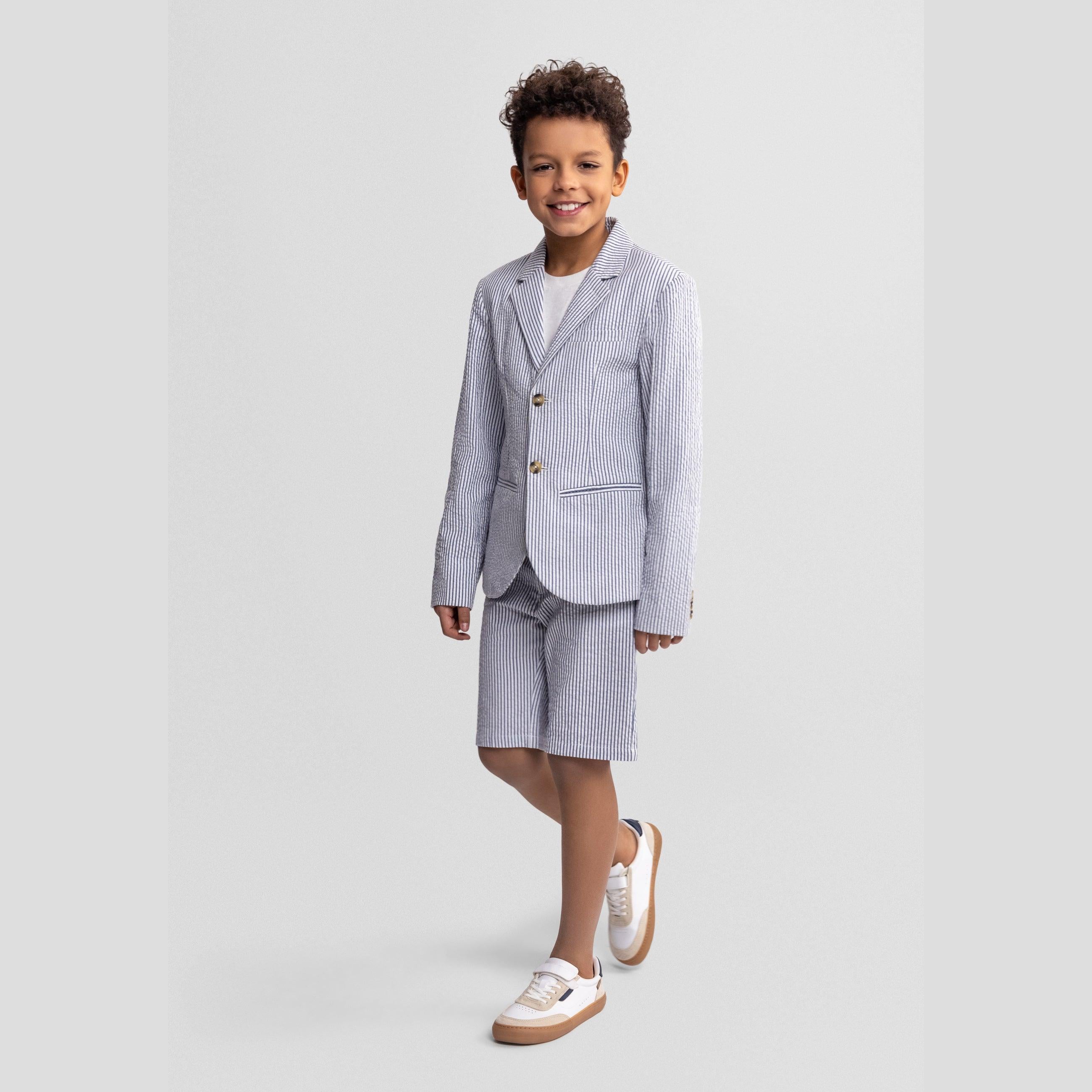 Full length front view of boy in navy striped blazer with white shirt and shorts, smiling naturally against neutral grey studio background