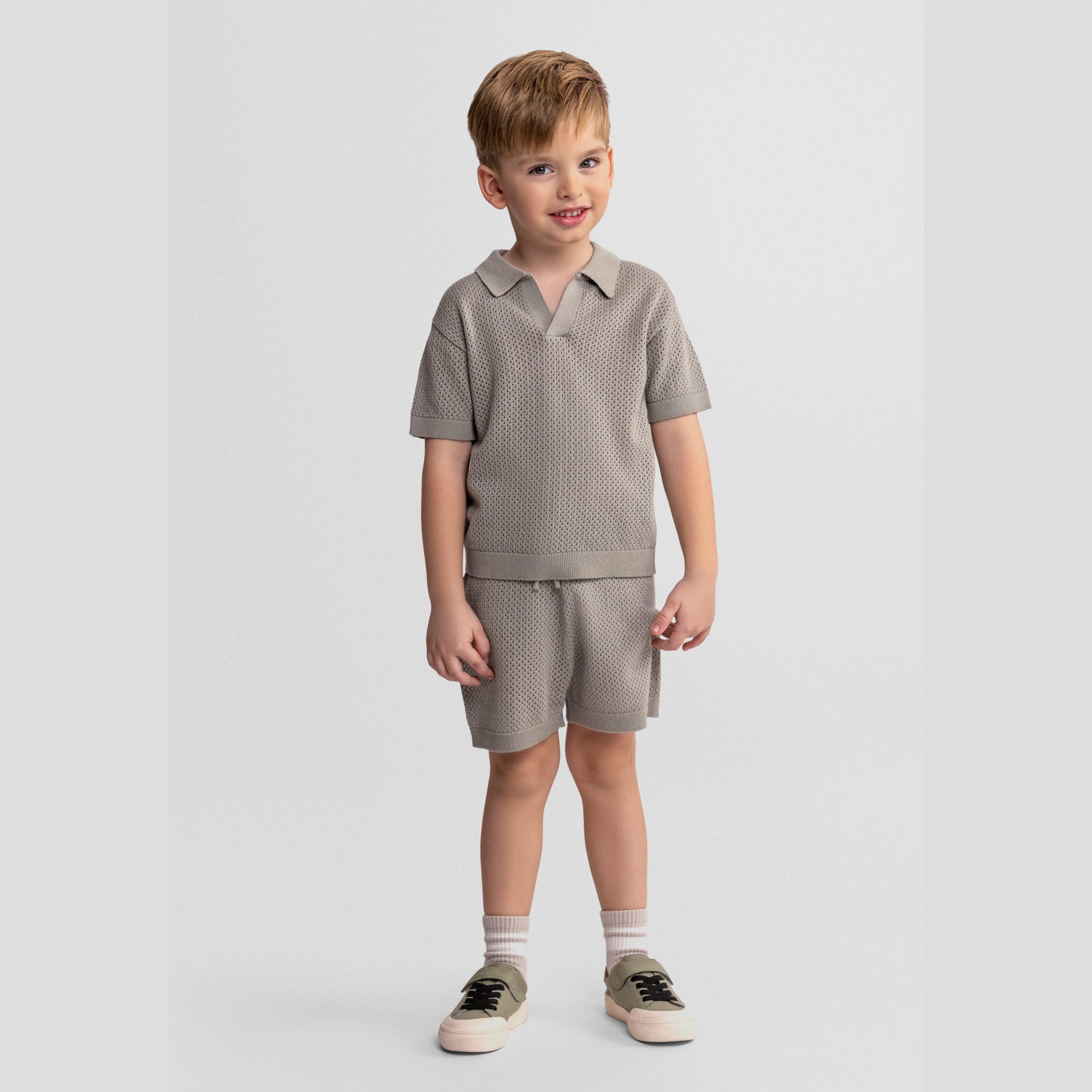 Young blonde boy wearing khaki knitted polo shirt and matching shorts set, standing on light grey background with hands by his sides, smiling at camera