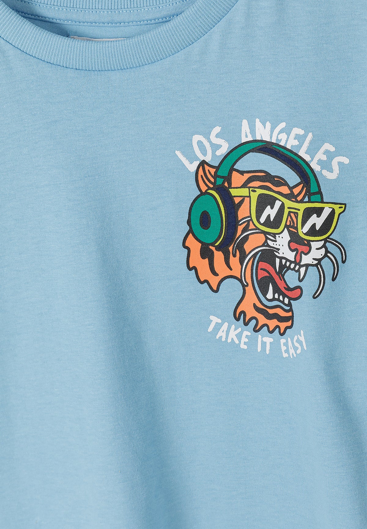 Close-up detail of light blue t-shirt showing colorful tiger head graphic with sunglasses, headphones and LOS ANGELES TAKE IT EASY text print on chest area