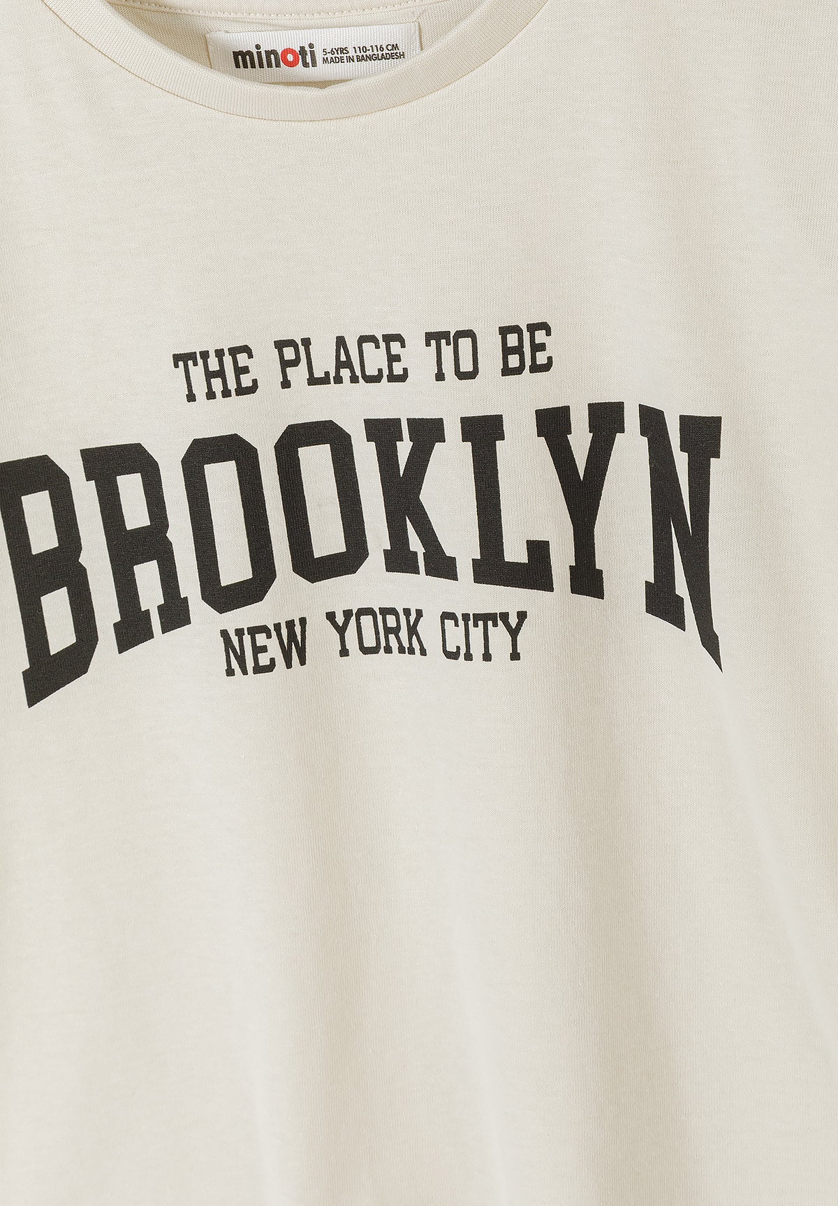 Close-up detail of cream t-shirt showing black The Place To Be Brooklyn New York City typography print with Minoti size label visible at neckline
