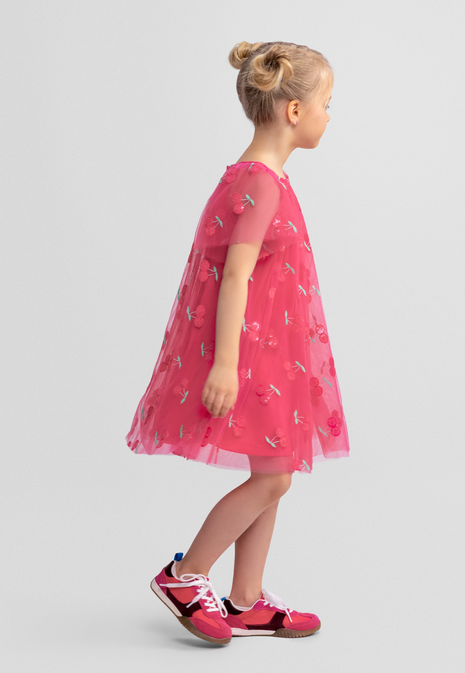 Side view of a young blonde girl wearing pink tulle dress with sequin cherry pattern and angel sleeves, standing against white background wearing pink shoes
