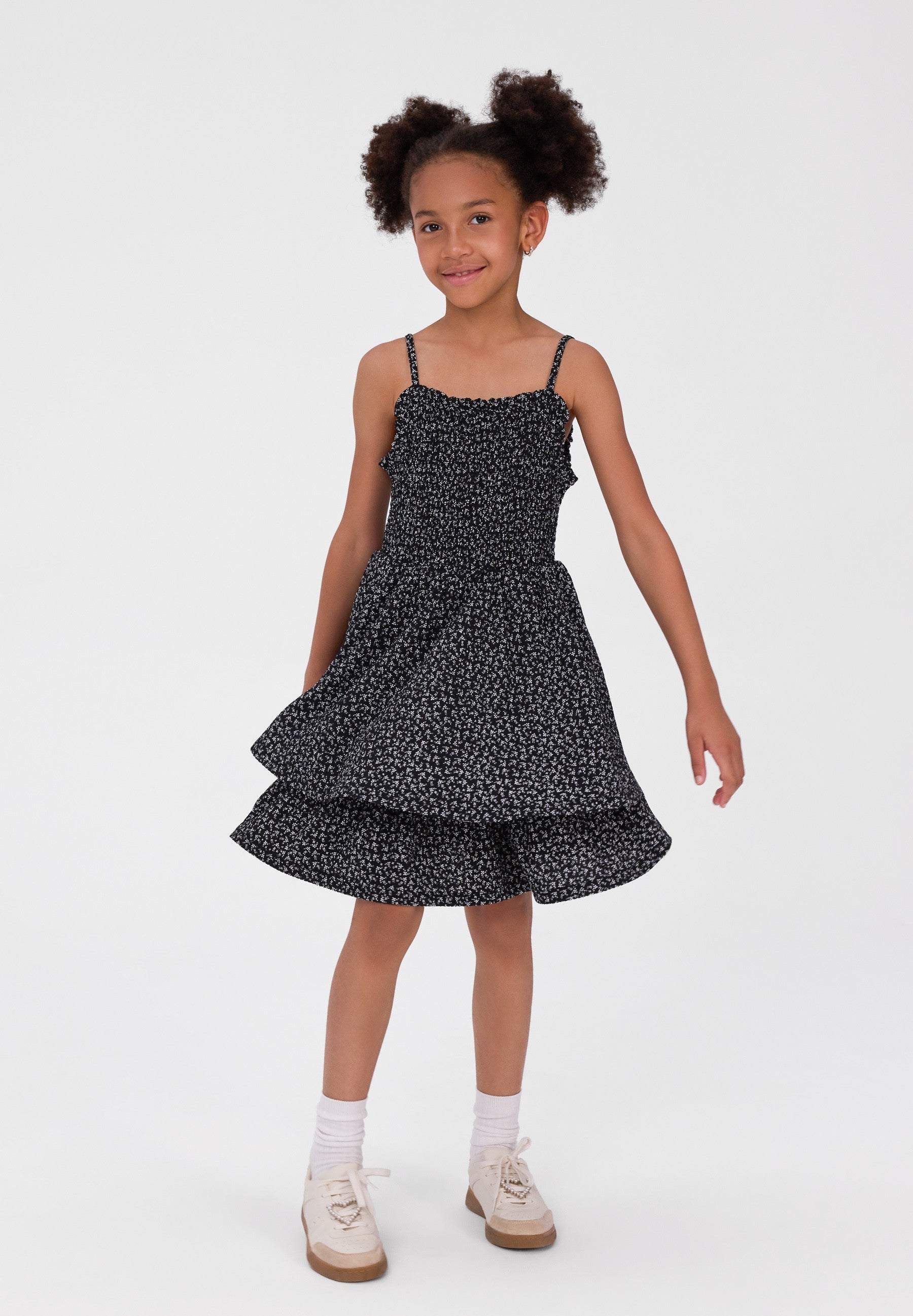 A young girl with curly hair is wearing a black and white micro-patterned spaghetti strap dress with a tiered skirt, paired with white socks and beige shoes, standing against a plain white studio background.