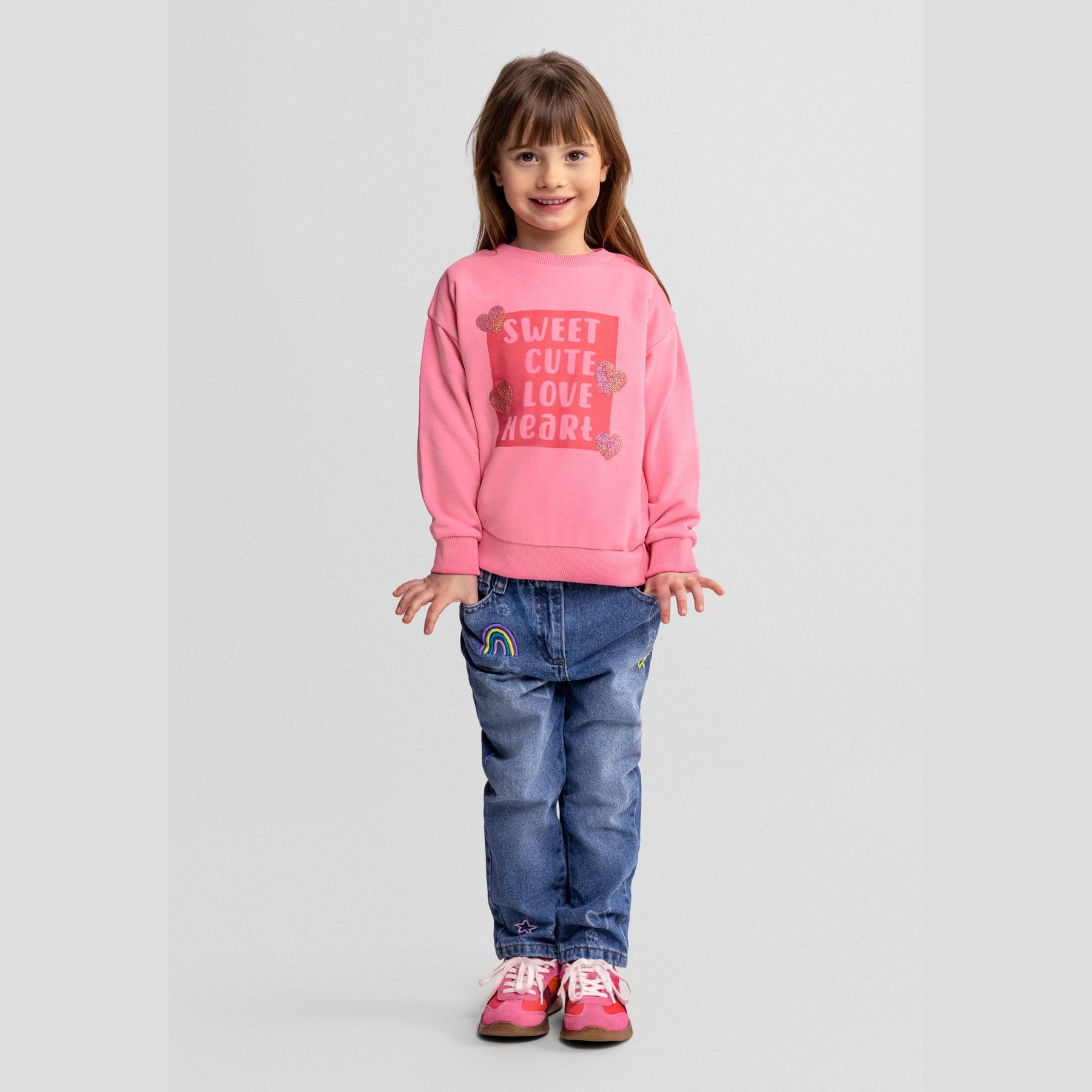 Young girl wearing pink drop shoulder sweatshirt with 'Sweet Cute Love Heart' print and sequined hearts, paired with denim jeans and pink sneakers, standing against light background