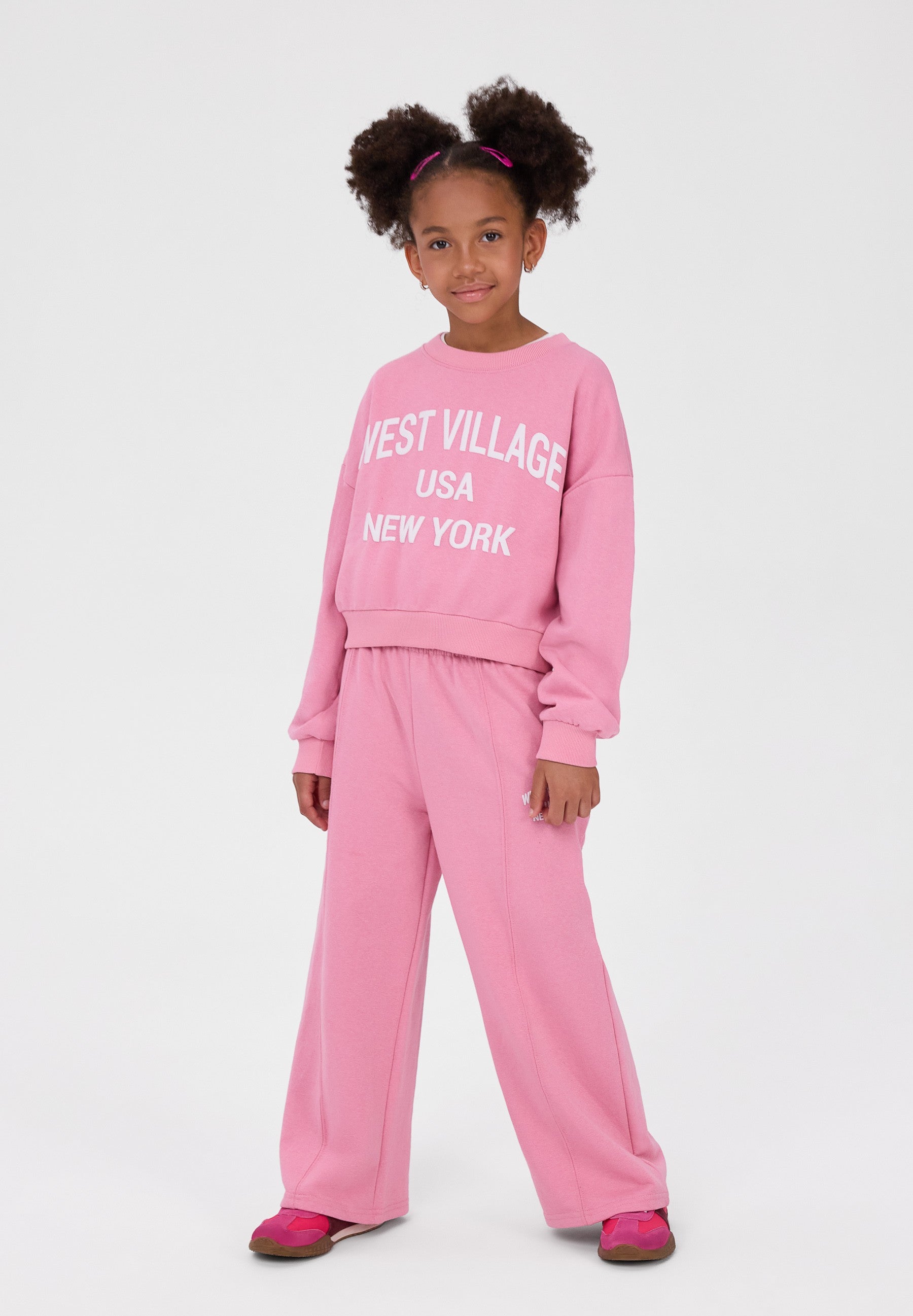 A young girl with two buns of hair wearing a pink two-piece sweatshirt and pants set with 'WEST VILLAGE USA NEW YORK' printed in white on the sweatshirt, standing against a light gray studio background.
