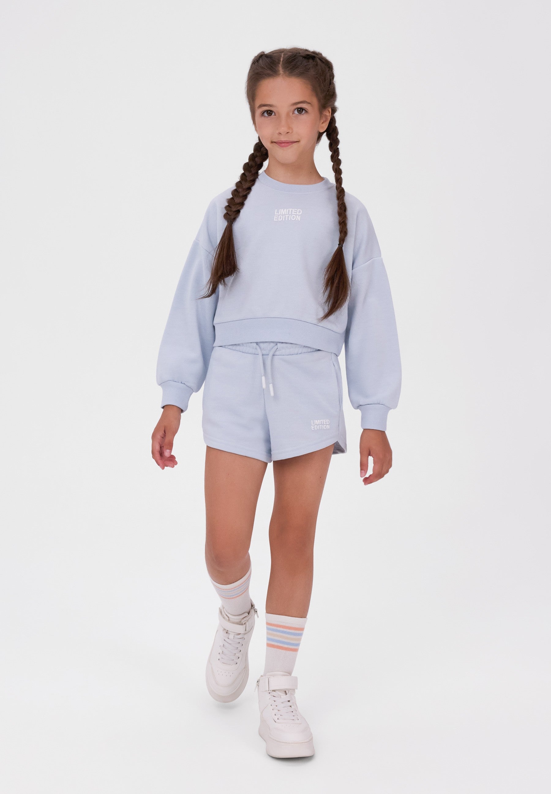 A young girl with braided hair models a light blue sweatshirt and matching shorts set, paired with white sneakers and striped socks, against a plain white studio background.