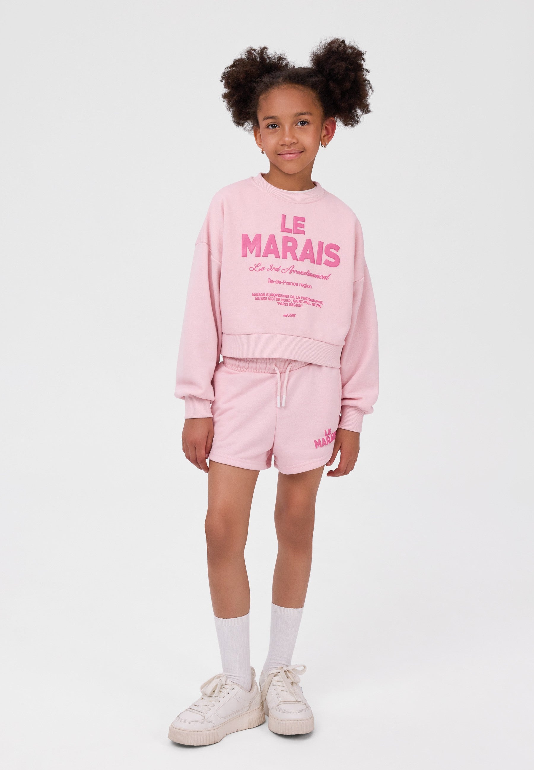 A young girl with curly hair styled in two buns stands against a plain white studio background wearing a light pink two-piece lounge set from Le Maïris. The set includes a cropped long-sleeve sweatshirt with pink text graphics on the front and matching drawstring shorts.