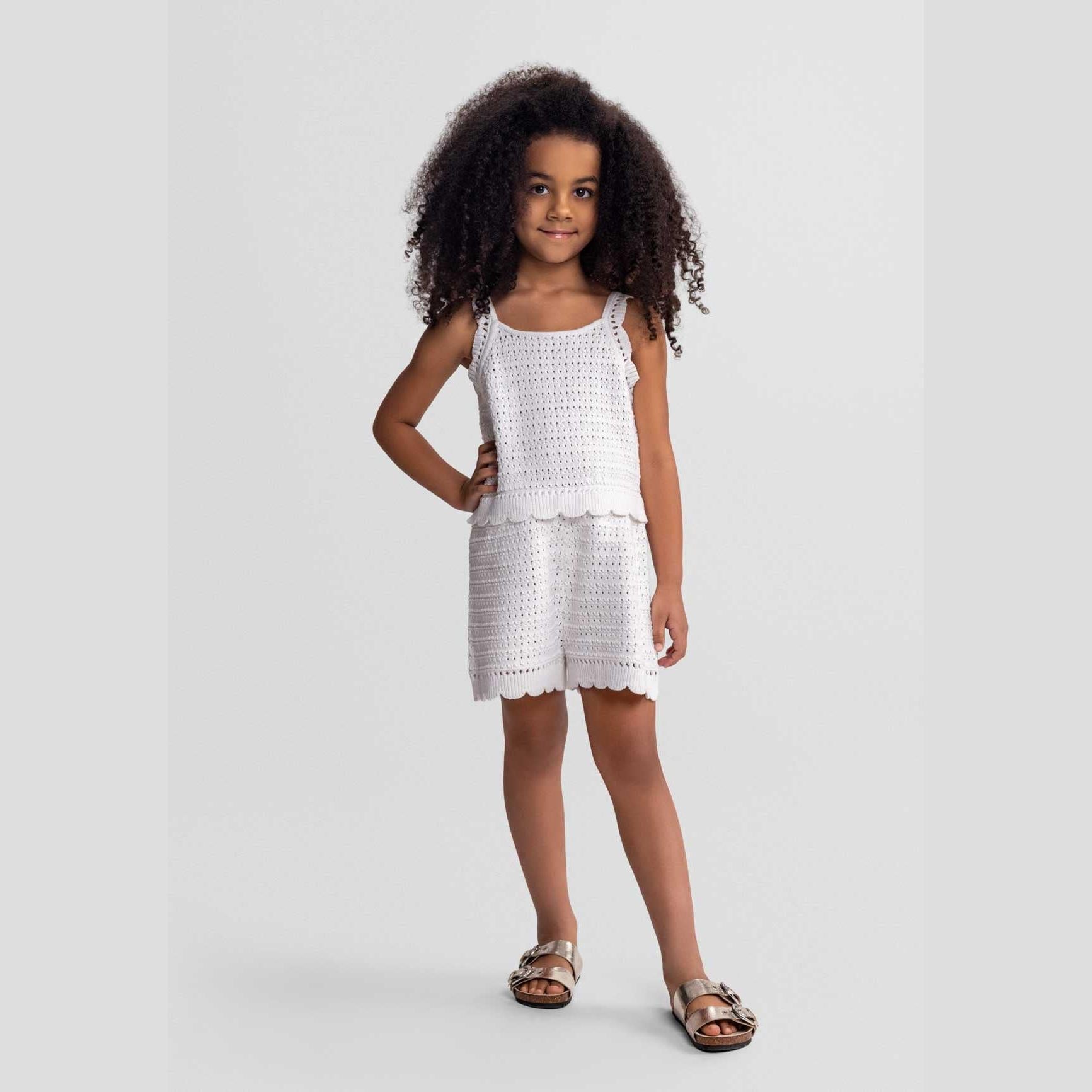 Young girl with curly hair wearing white pointelle knit romper with wide straps and scalloped hem, paired with brown sandals against grey background