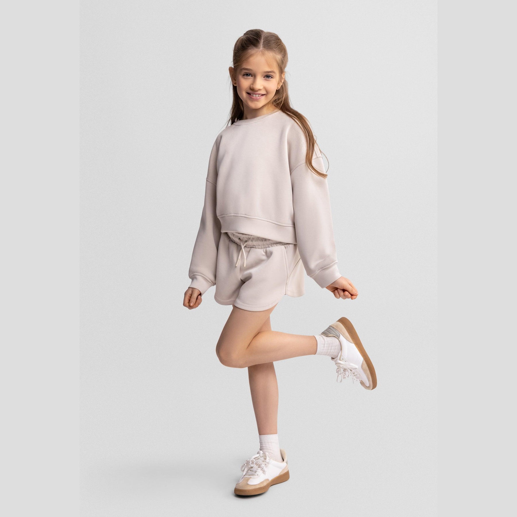 Young girl wearing beige sweatshirt and shorts set, posing playfully on one leg against light grey background