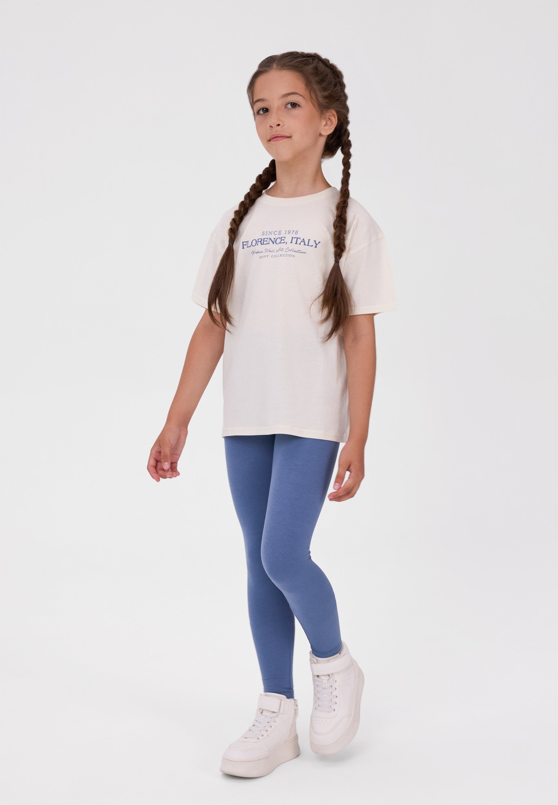 A young girl with braided hair stands against a light gray studio background wearing a white graphic t-shirt, blue leggings, and white sneakers. The t-shirt features a blue graphic print that reads 'COSTUME D'ORLANDO, FLORIDA, ITALY'.