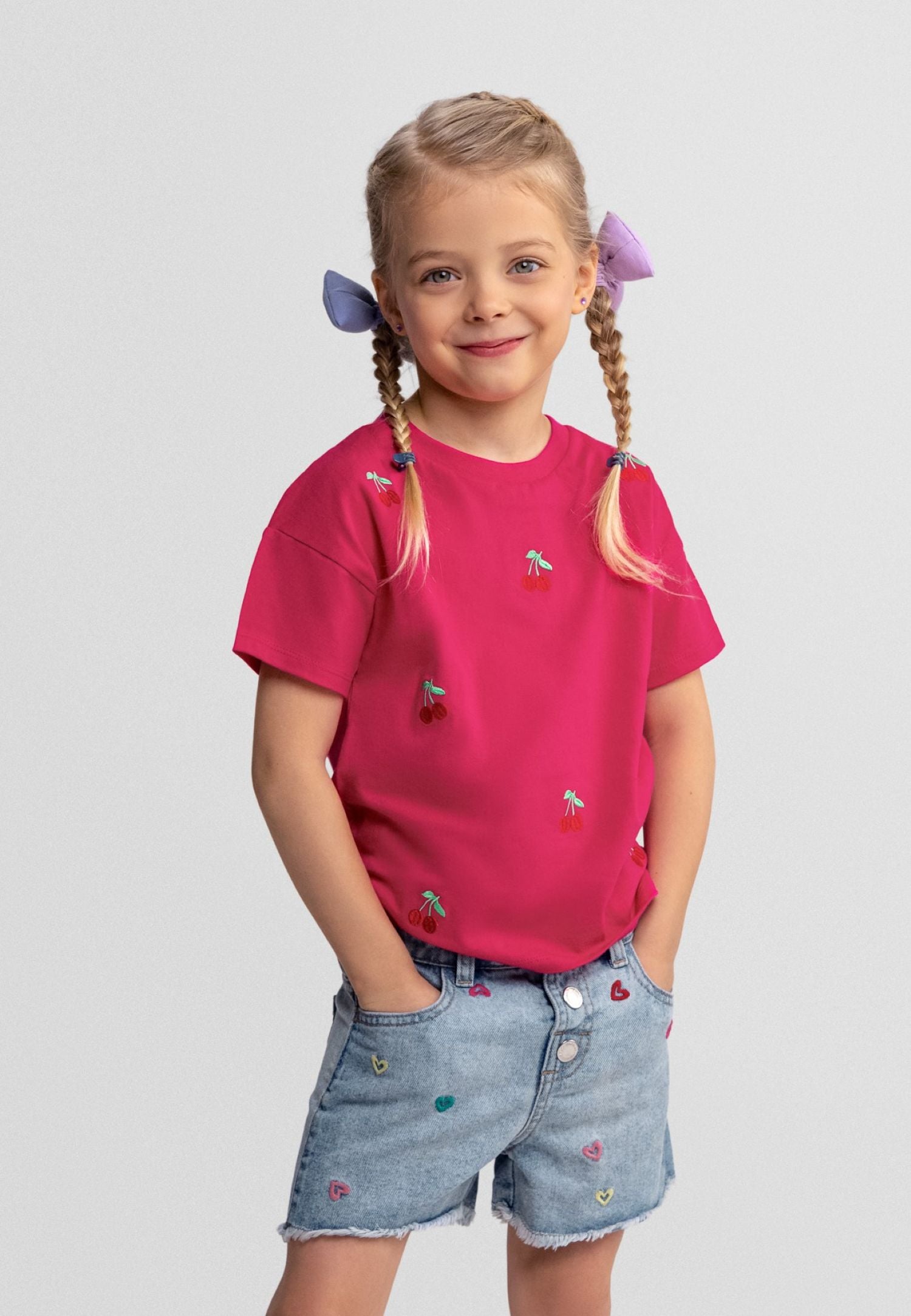 Young girl wearing a pink shirt and denim shorts with colorful buttons on a light gray background