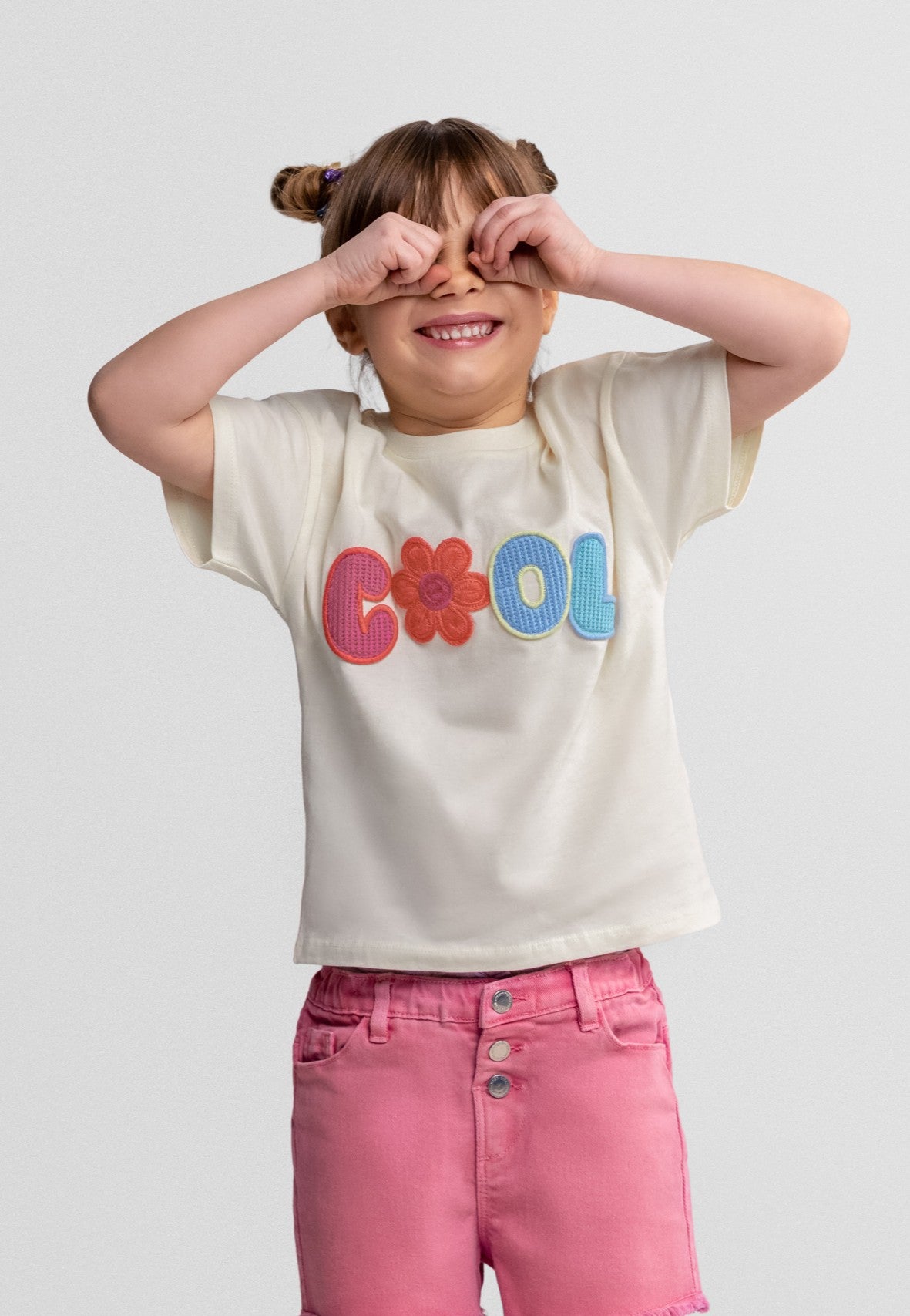 Child wearing a white t-shirt with colorful letters and pink pants on a light gray background