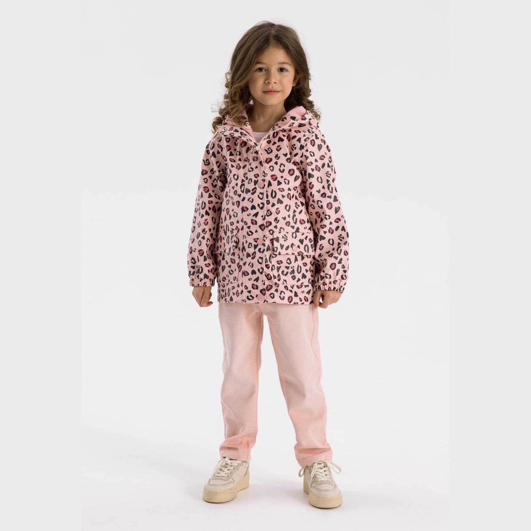Young girl wearing pink leopard print hooded rain jacket with matching pink trousers and beige trainers, standing against white background