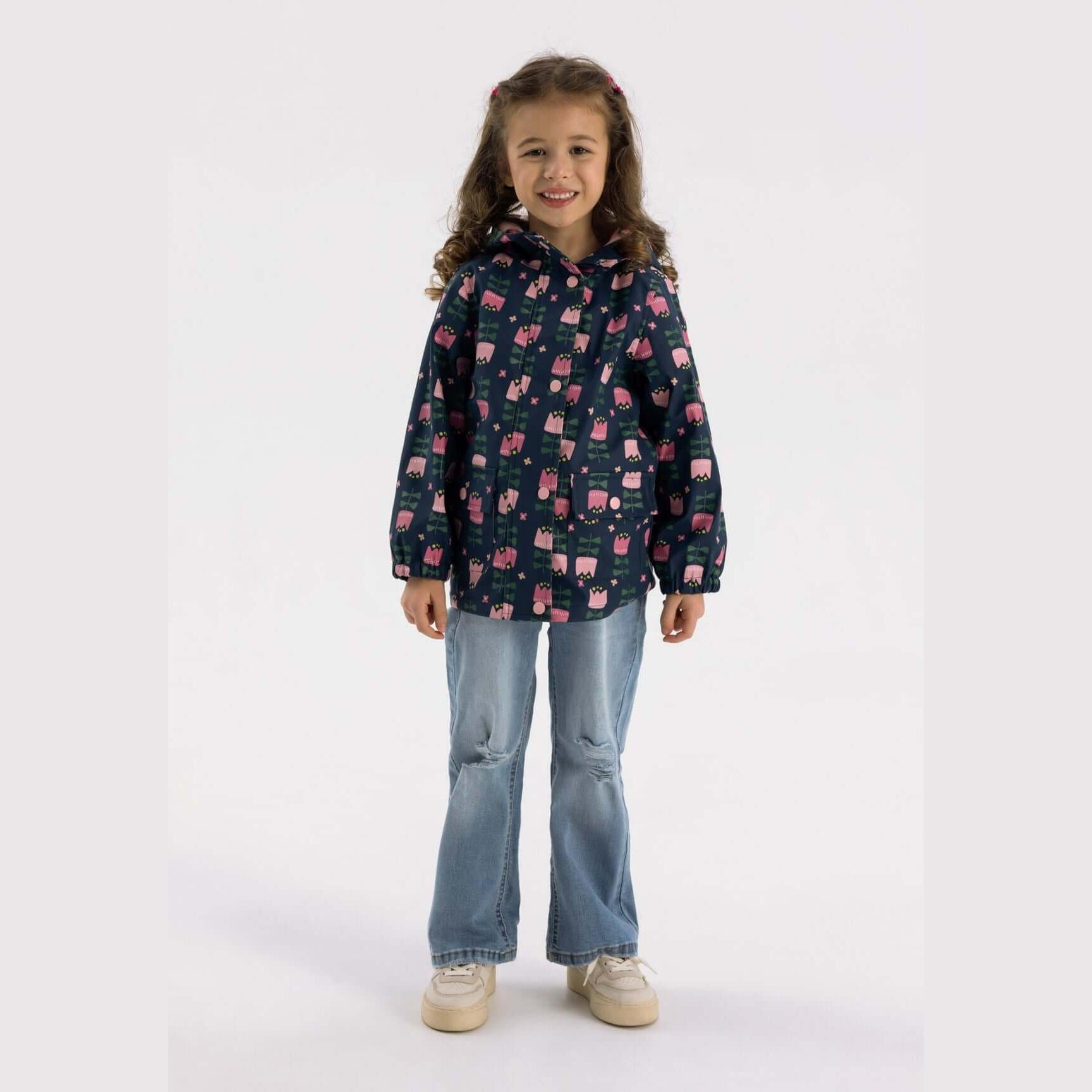 Young girl wearing navy rain jacket with pink tulip print and light blue jeans, standing against white background