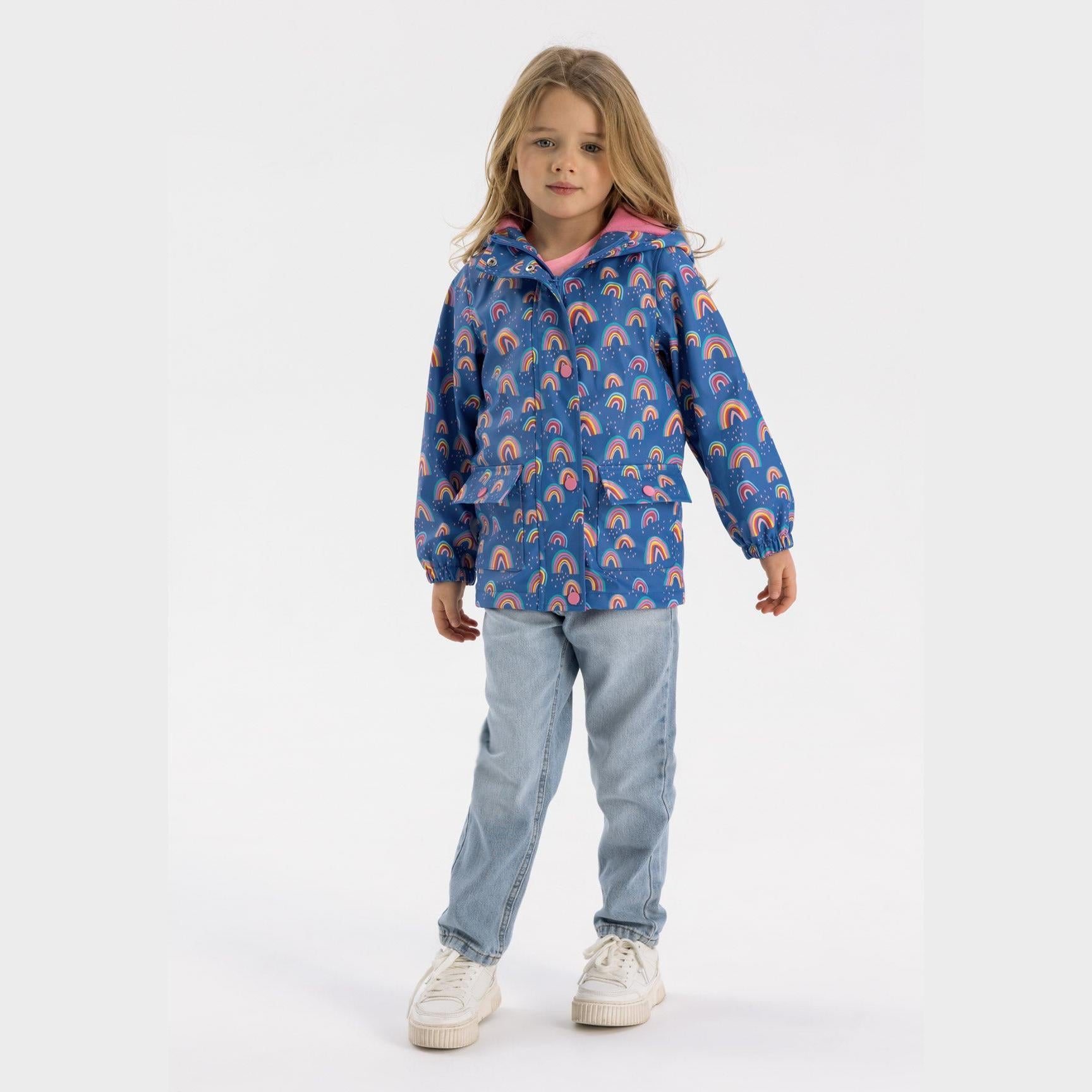 Young girl wearing cobalt blue hooded rain jacket with rainbow print, paired with light wash jeans and white sneakers on white background