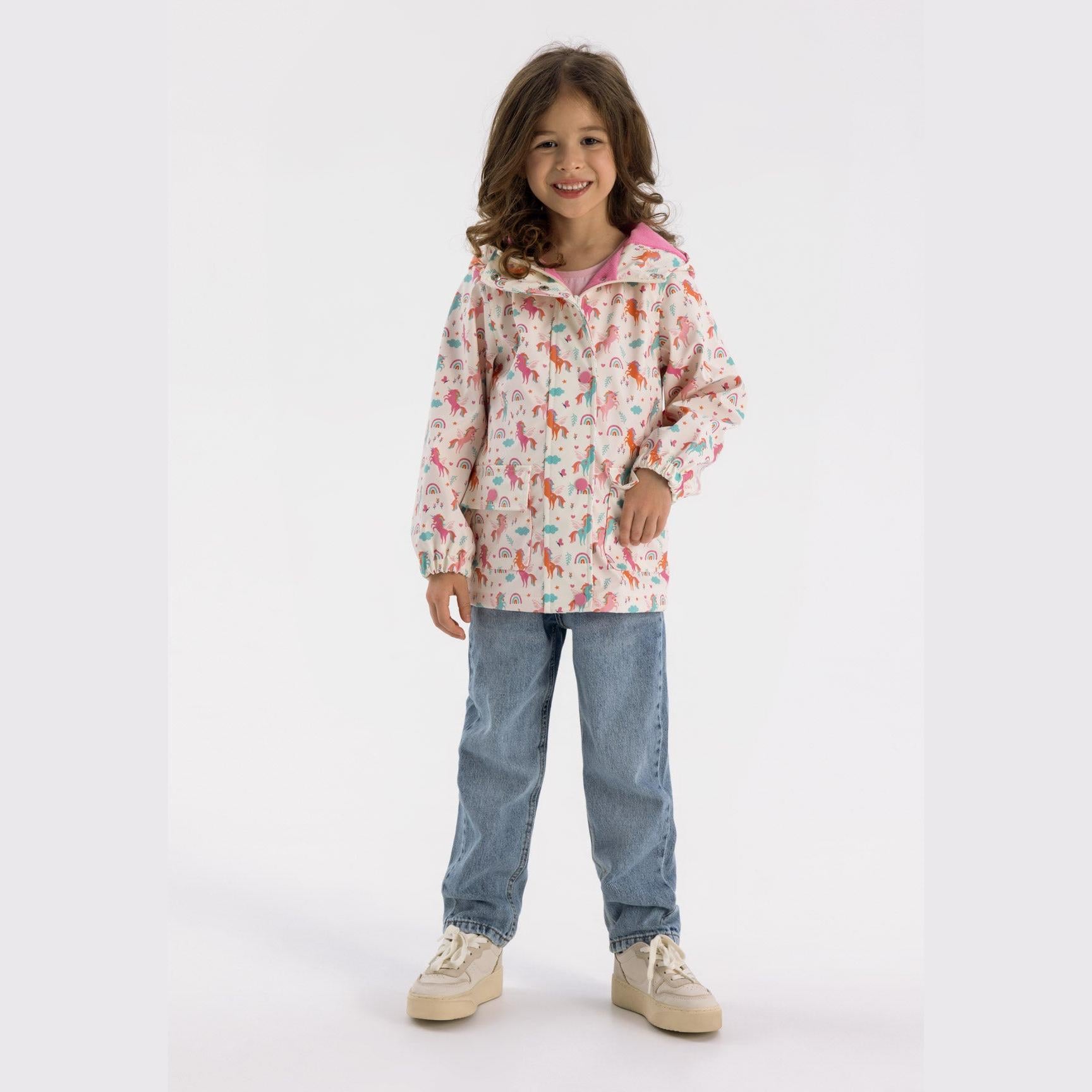 Young girl wearing cream unicorn print hooded rain jacket with jeans and beige sneakers against white background