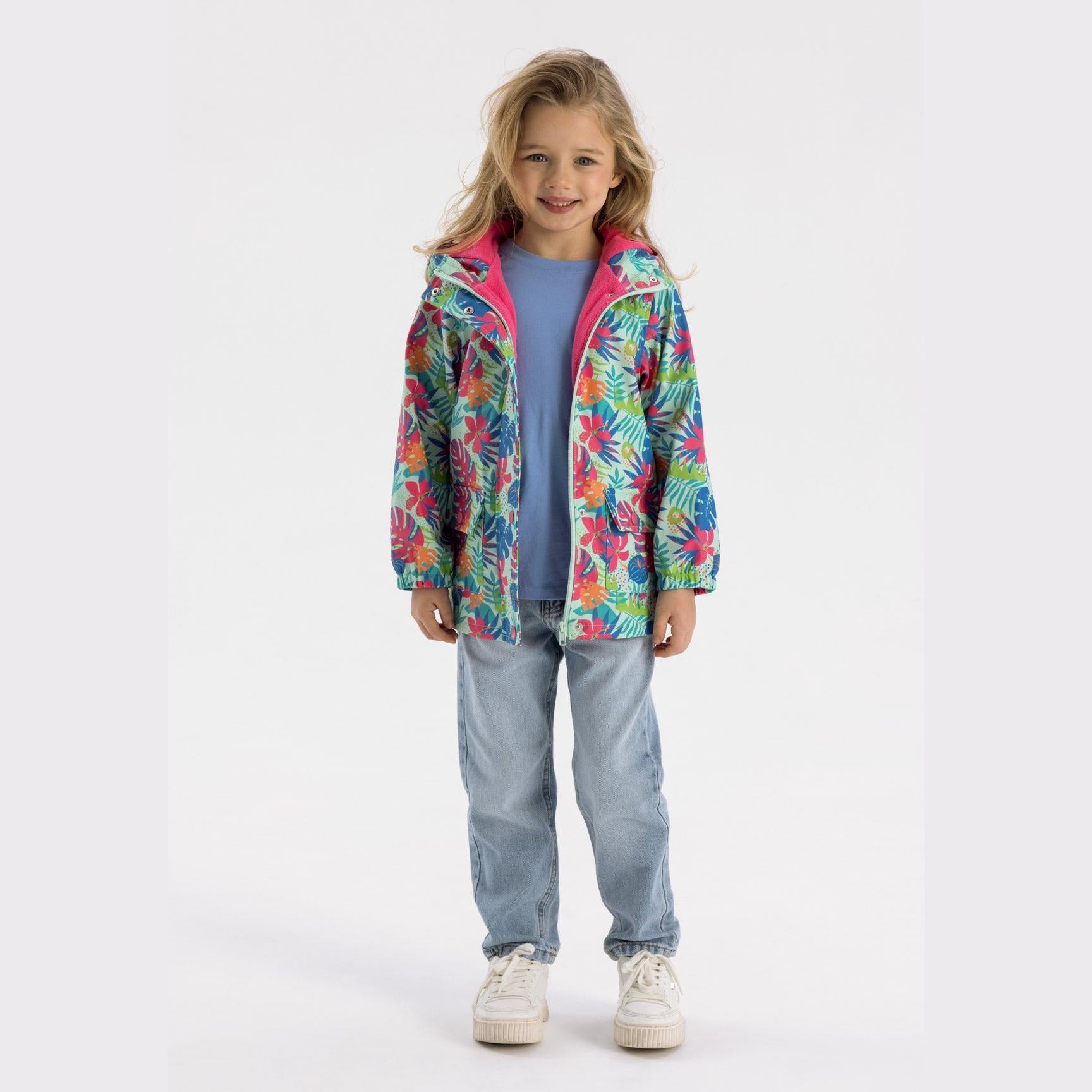 Young girl wearing tropical print rain jacket in mint green with hood down, paired with light blue top and denim jeans, standing against white background