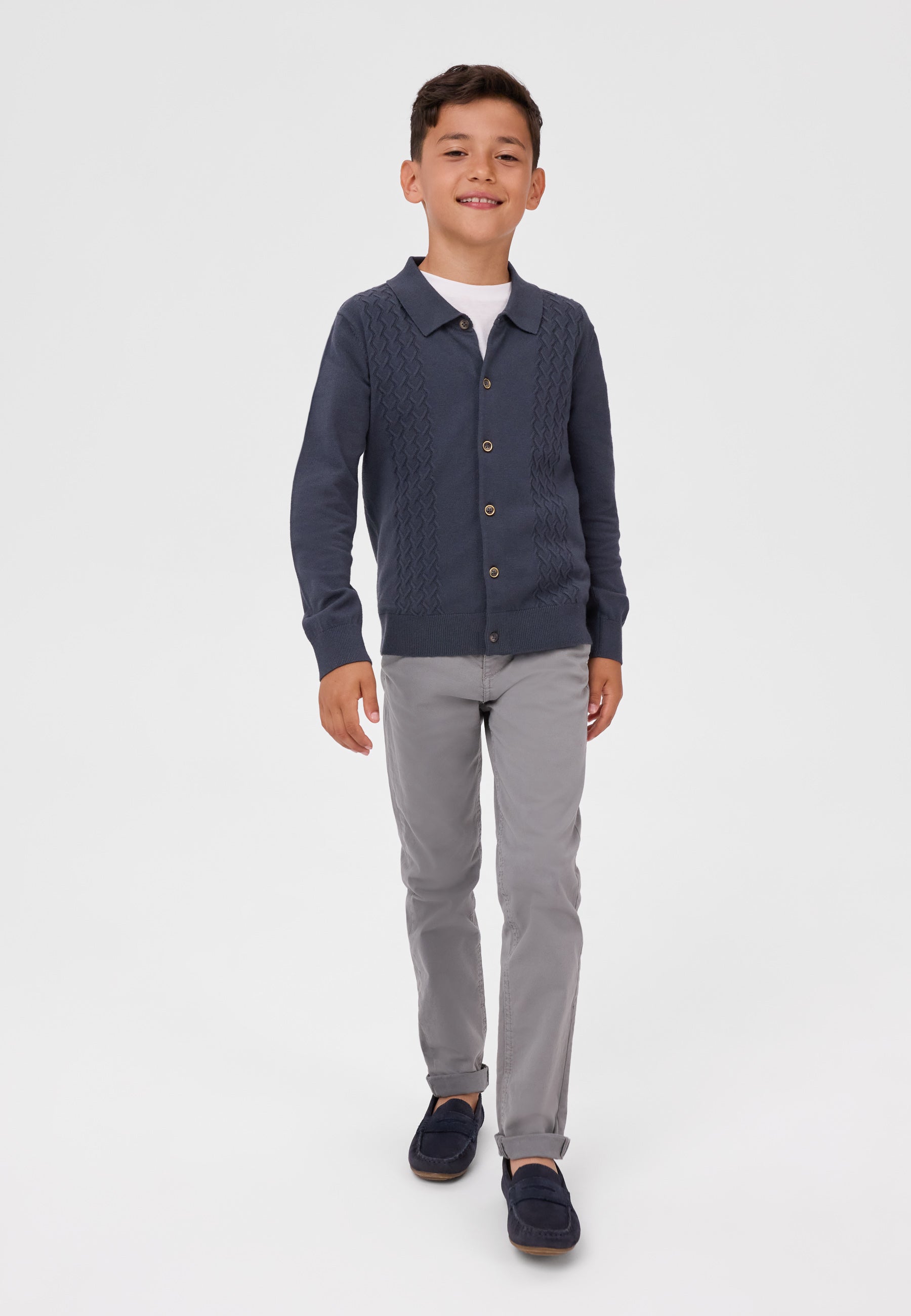 A young boy modeling a navy blue cable-knit button-up cardigan over a white t-shirt, paired with gray trousers and navy loafers, on a light gray studio background.