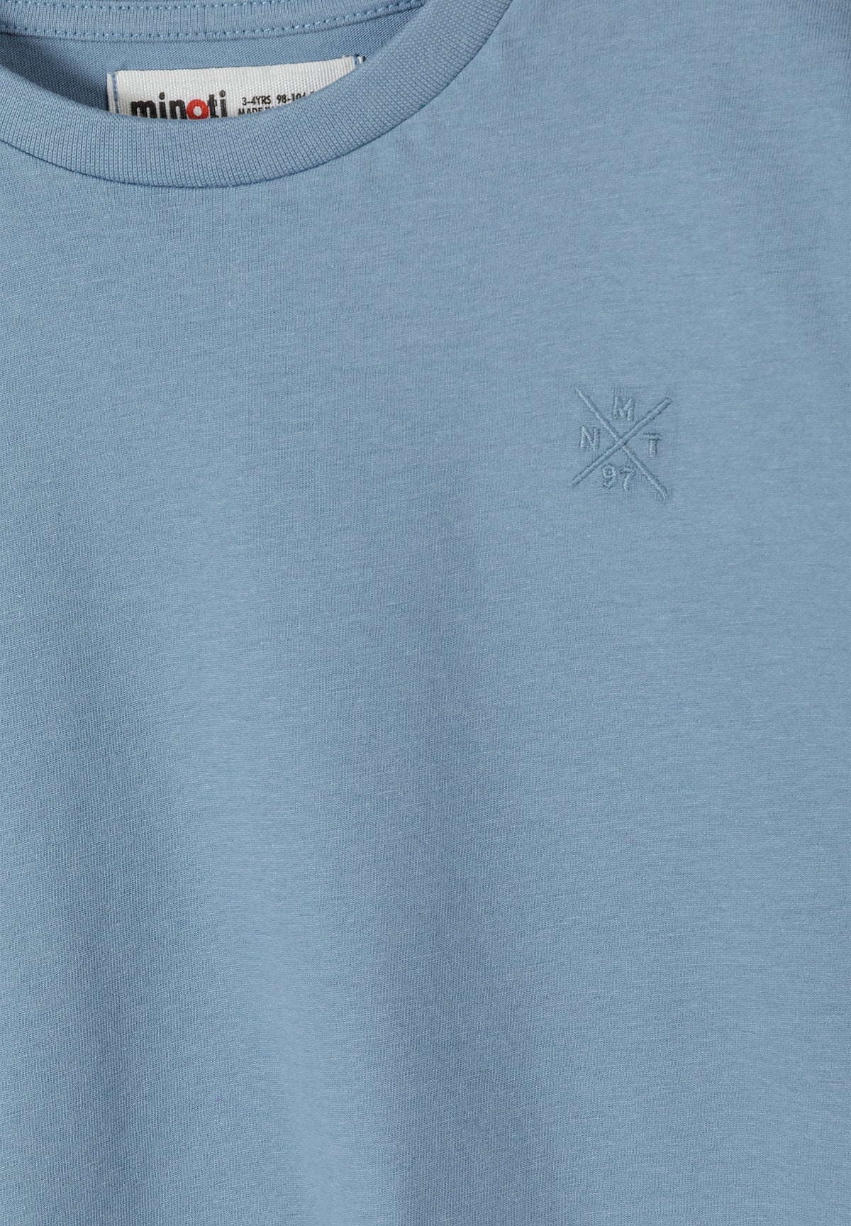 Close-up detail of dusty blue Minoti t-shirt showing crew neckline with brand label and small embroidered cross logo on chest