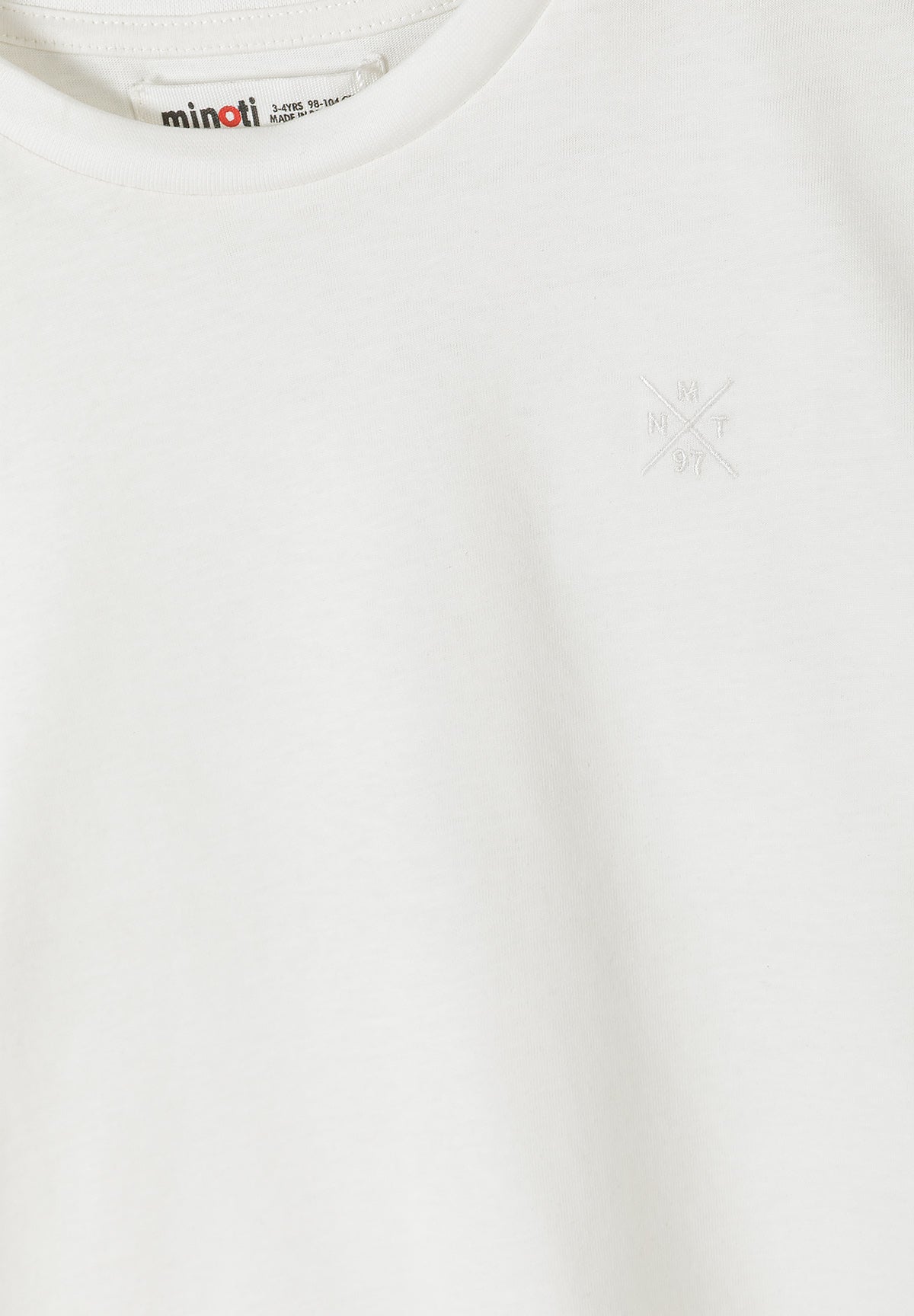 Close-up detail of white cotton t-shirt showing Minoti brand label at neckline and small embroidered cross logo on chest area