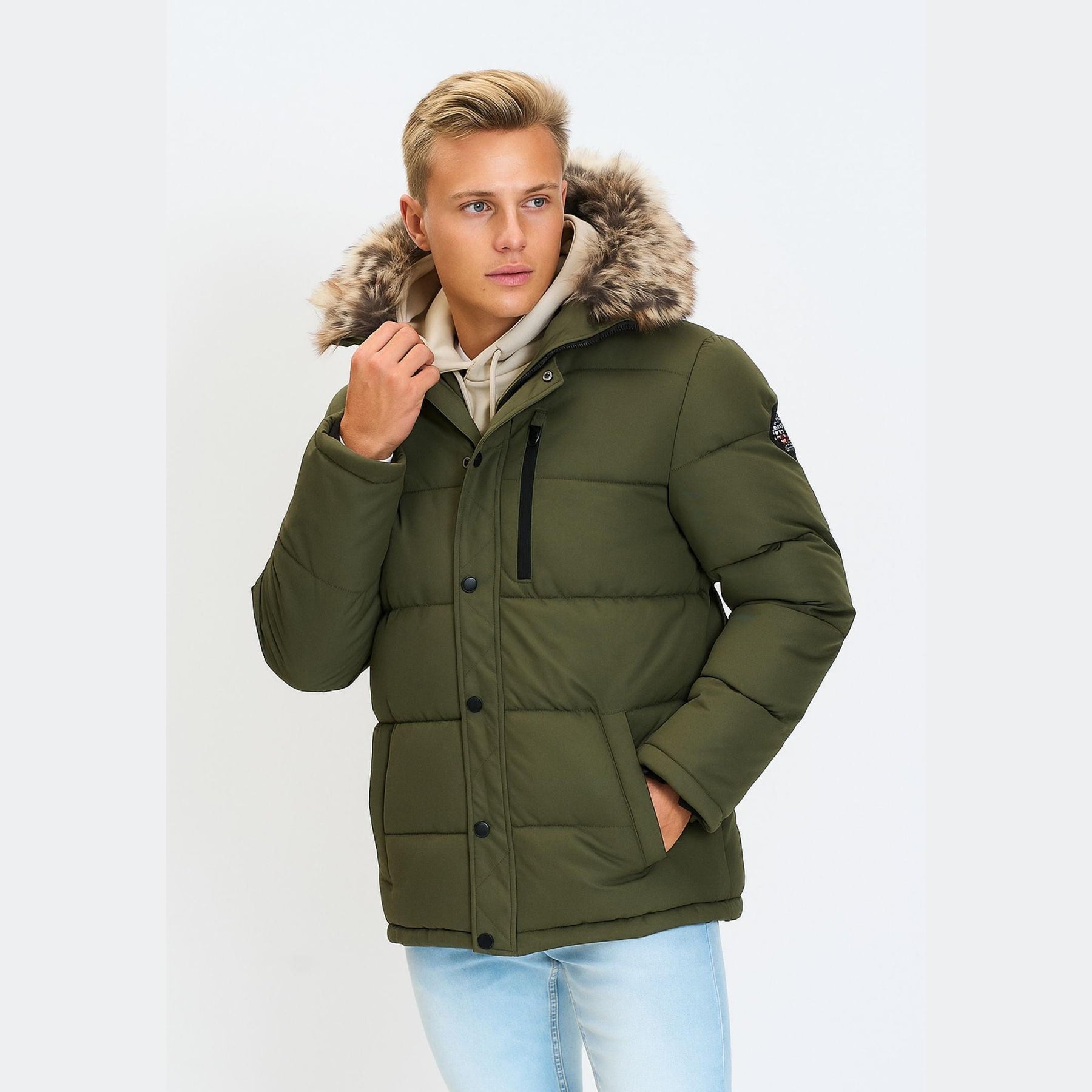 Young male model wearing khaki padded jacket with faux fur hood over cream hoodie and light blue jeans, standing against white background