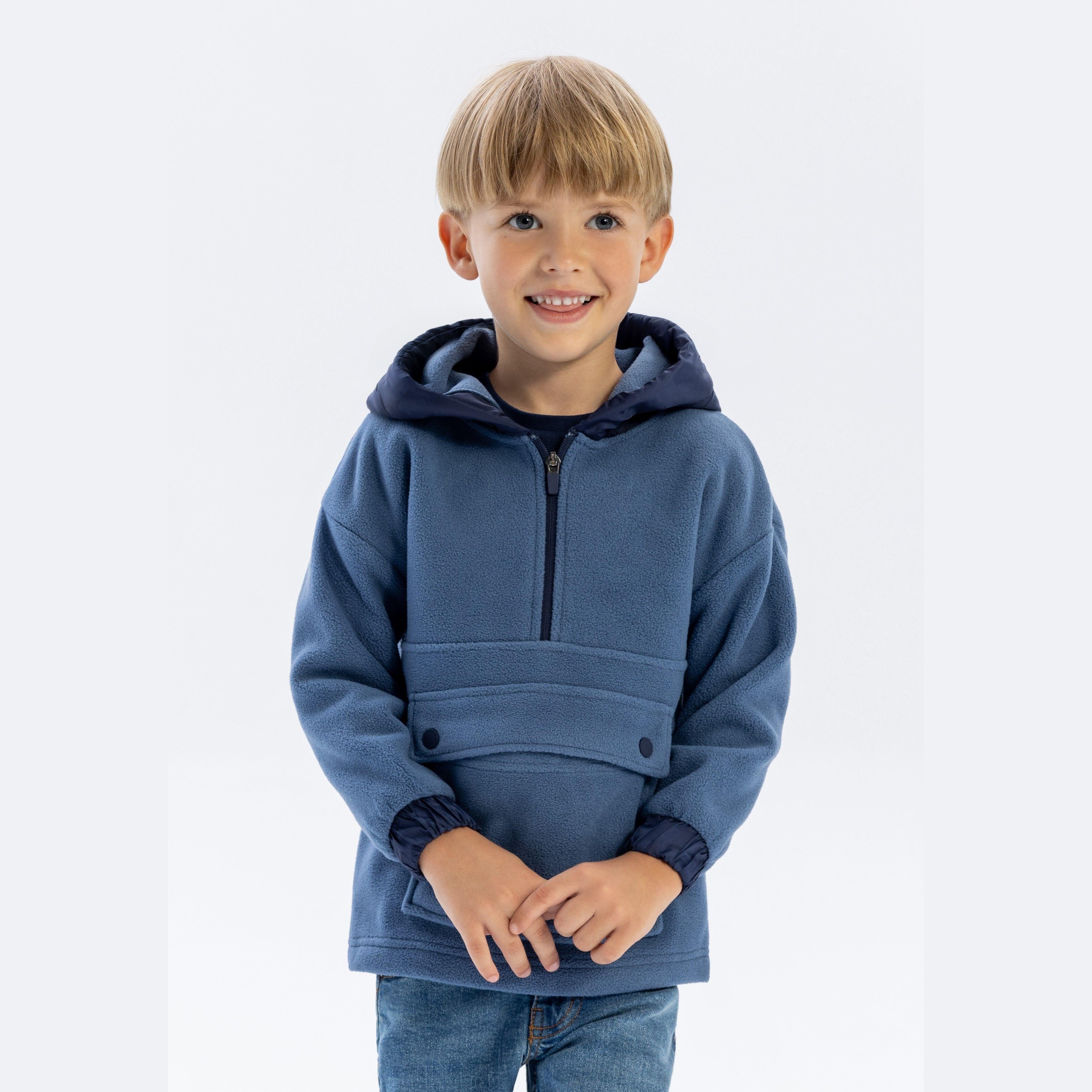 Young blonde boy wearing blue microfleece half-zip hoodie with hands clasped, smiling at camera against white background
