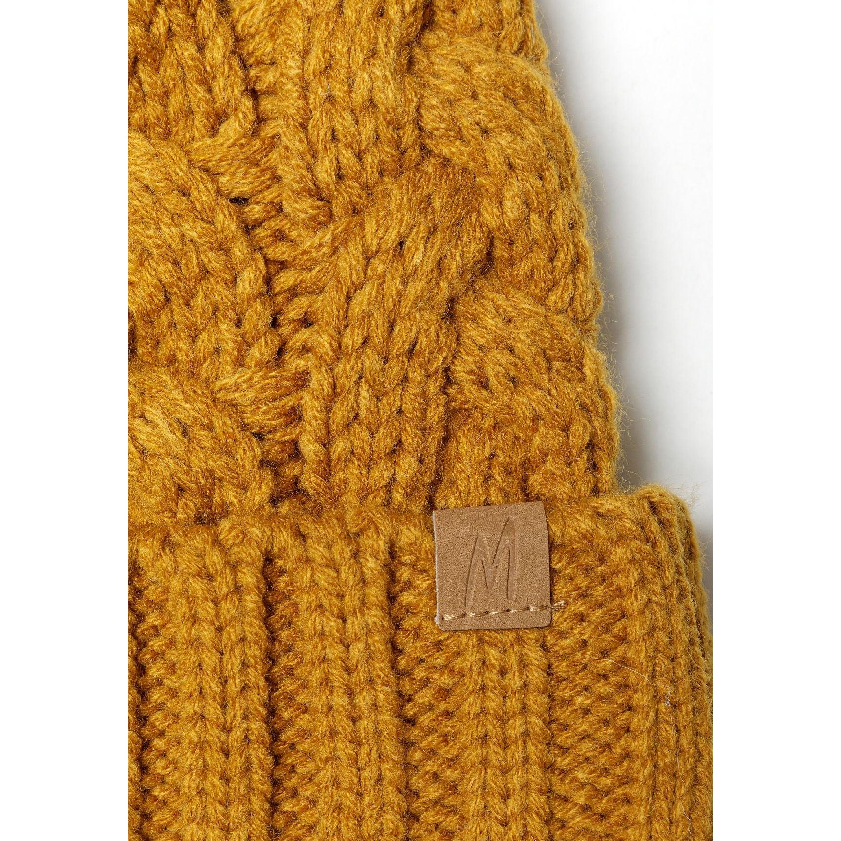 Close-up detail of mustard cable knit beanie showing intricate twisted cable pattern texture and brown leather tab with embossed M logo