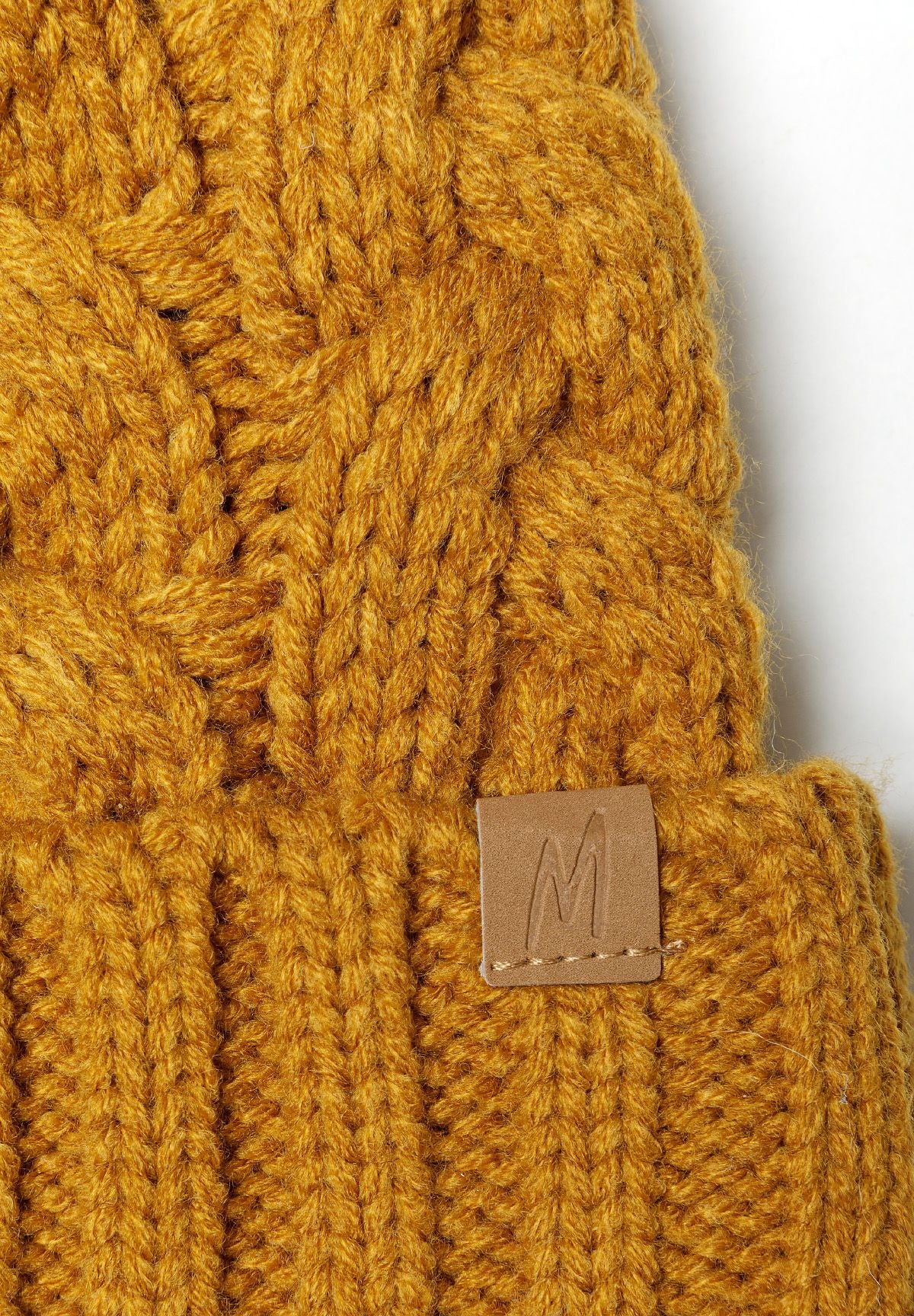 Close-up of a mustard knitted cable hat with a pom pom and a small tab detail.
