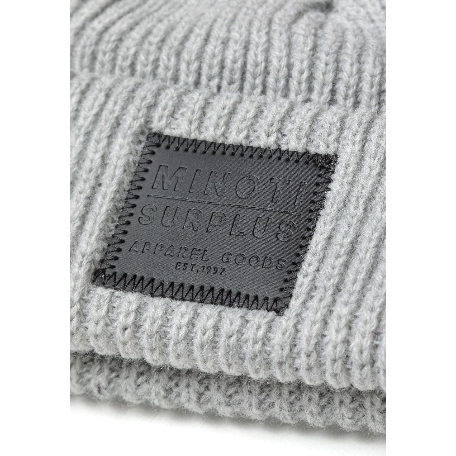 Close-up detail of dark grey rectangular leather patch on light grey ribbed beanie showing Minoti Surplus Apparel Goods Est. 1997 text with decorative stitching