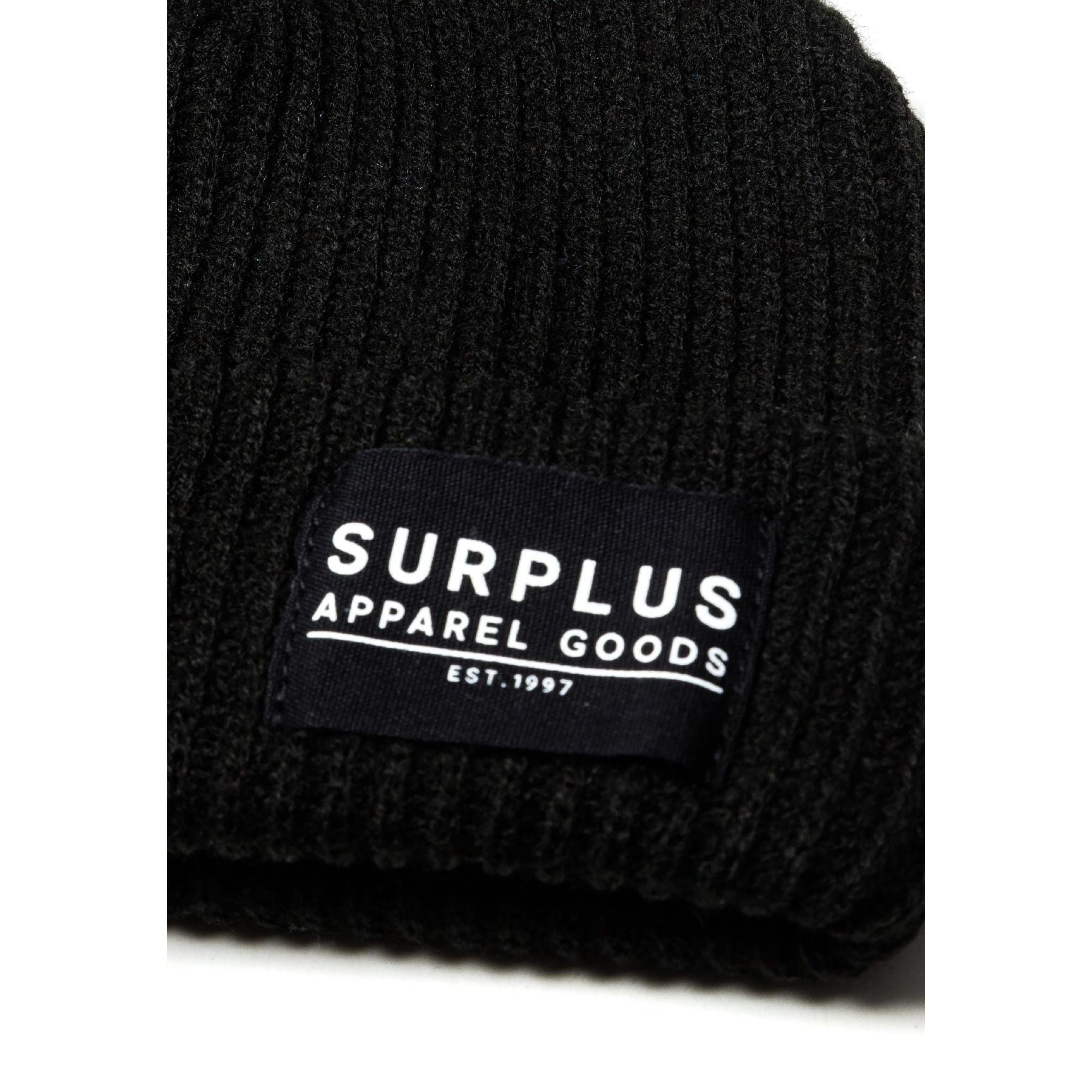 Close-up detail of black beanie showing SURPLUS APPAREL GOODS EST. 1997 woven label on ribbed knit fabric